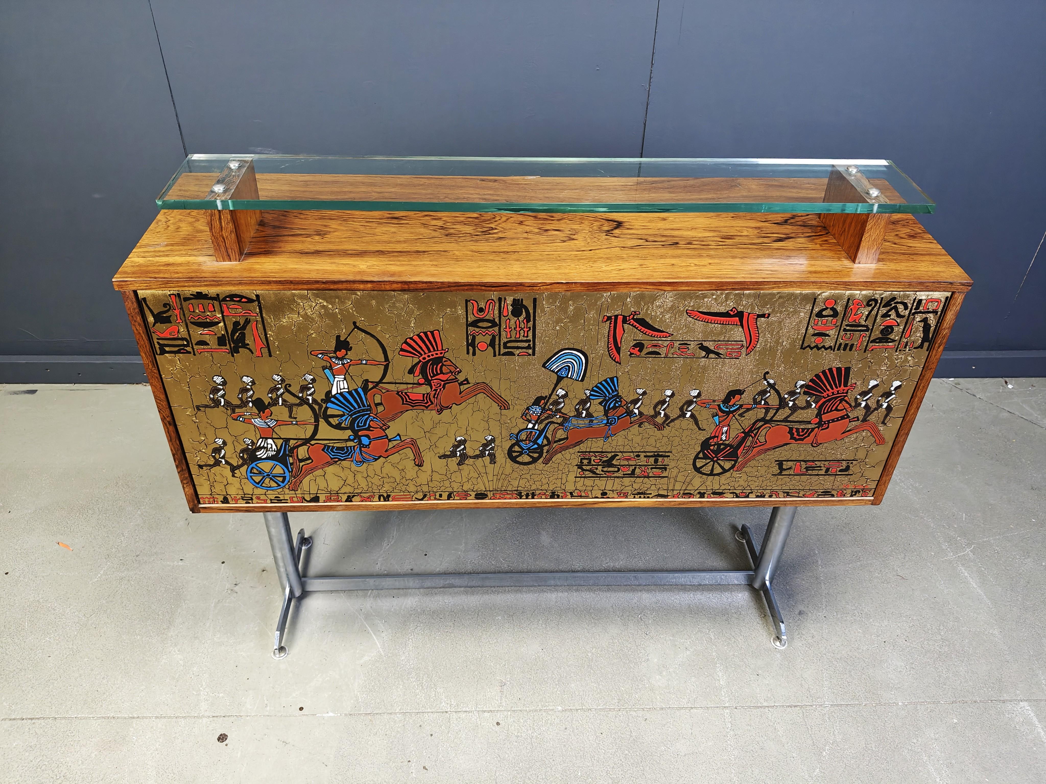 
Mid century bar counter by Denisco featuring a chromed metal base, a wooden case with a small glass top and a painted egyptian scene.

beautiful mid century piece.

Signed bottom right

On the inside we will provide some glass shelves or you can