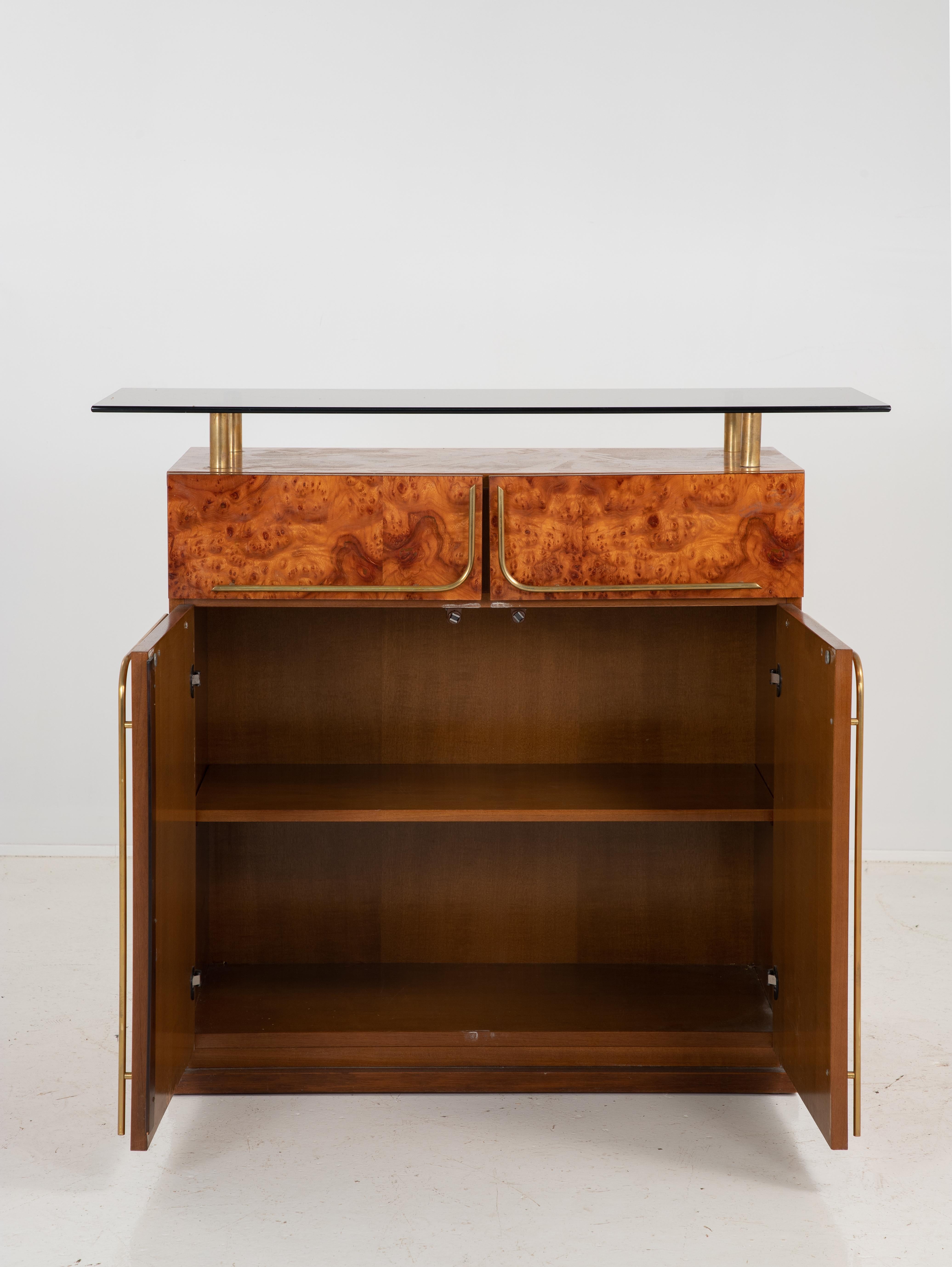 A mid century bar with brass handles and smoked glass elevated shelf. There are two drawers and the door cabinet opens up to reveal a shelf. The top of bar has four brass columns supporting a smoked glass shelf that extends beyond the width of the