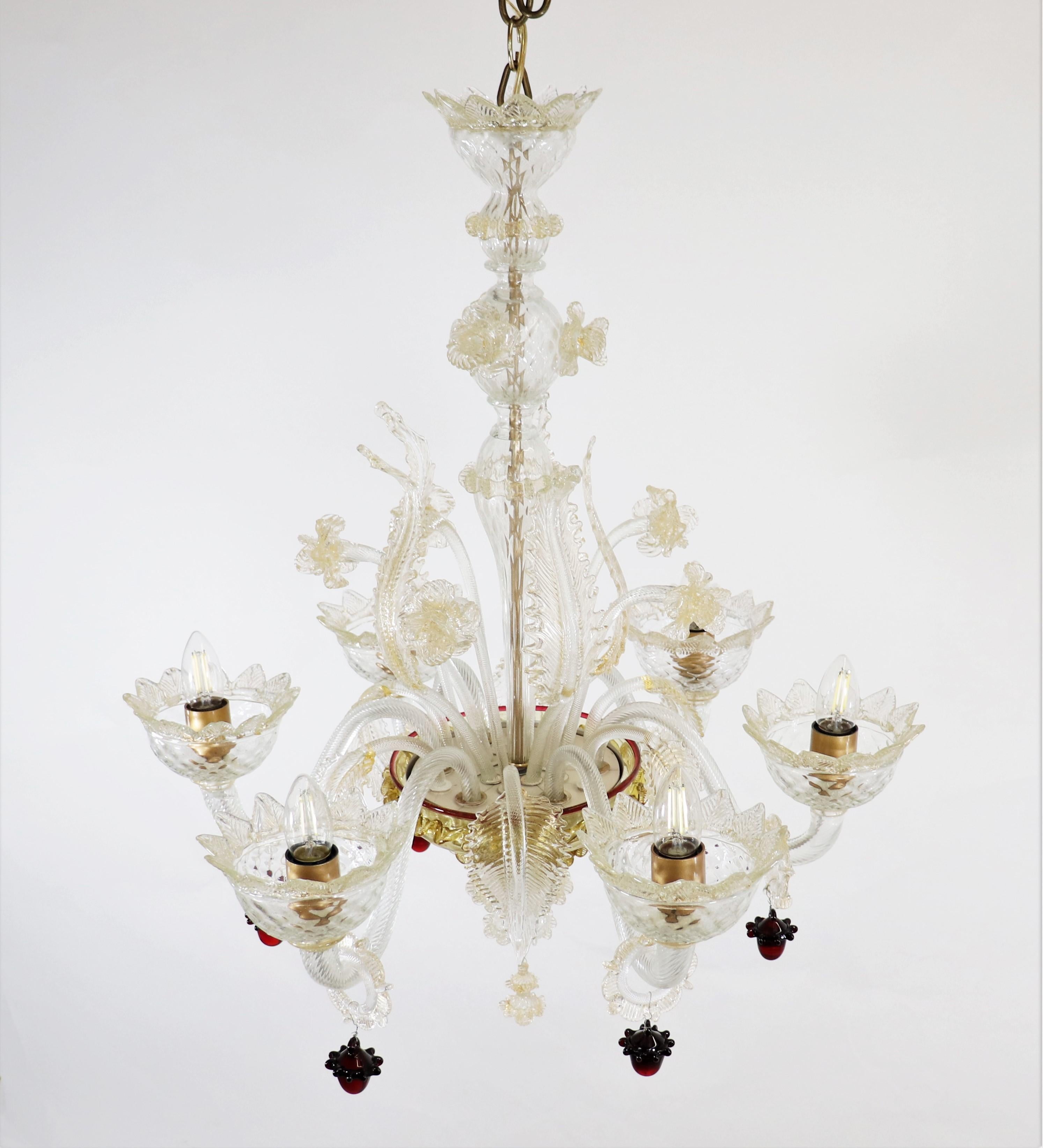 Questo lampadario di Murano, squisitamente realizzato a mano in cristallo infuso d'oro, ha sei bracci ondulati a voluta con baubles pendenti di colore scarlatto, che si abbinano alle finiture rosse della ciotola centrale. Foglie di vetro e narcisi