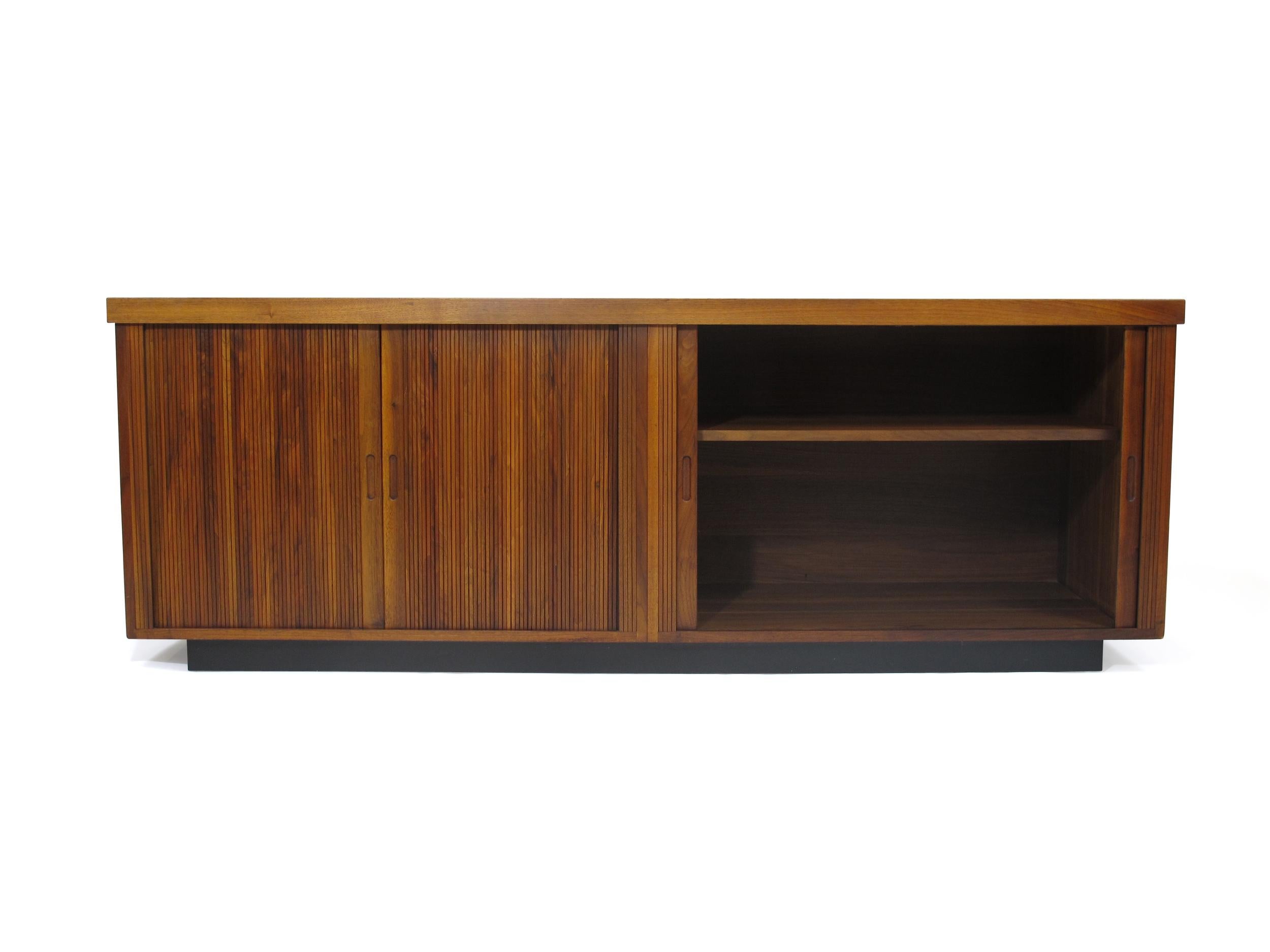 Midcentury Barzilay walnut credenza with pair of tambour doors with inset pulls which open to reveal an interior with adjustable shelves. Raised on a plinth base with black Formica top surface. Very good original condition.