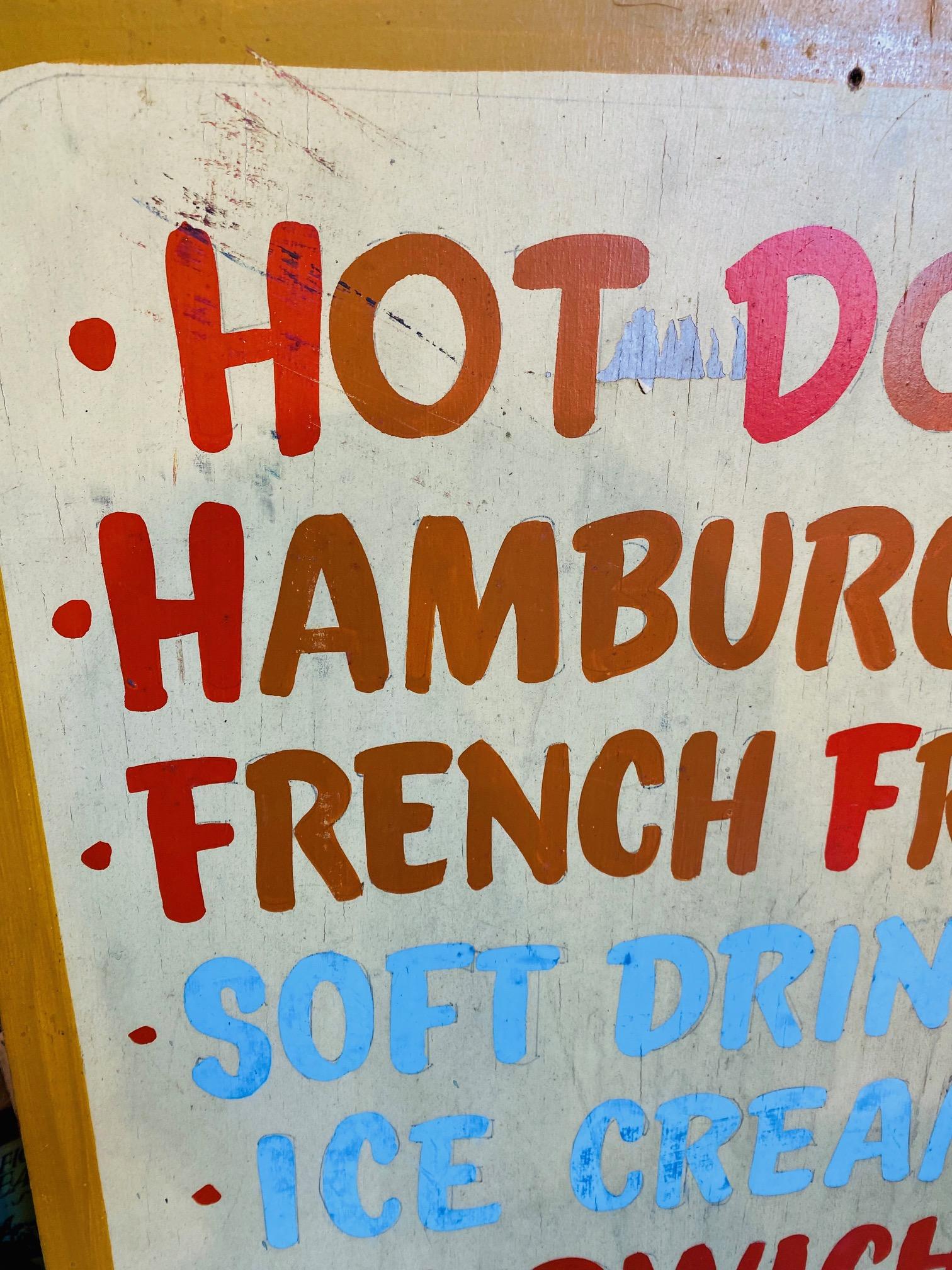 Vintage mid century beach-side hamburger shack menu board, circa 1950s, hand painted on plywood advertising hot dogs and hamburgers and the usual suspects... plus sunglasses. And Bathers are welcome!

Sign remains in very good weathered condition.