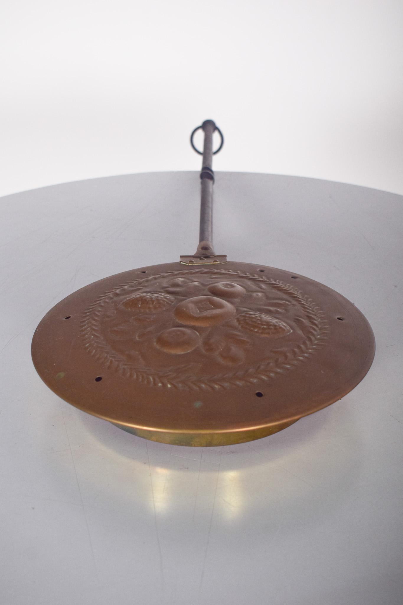 Brasero ancien avec poignée. L'objet se compose d'un récipient métallique en laiton avec un couvercle décoré en relief de motifs floraux et fruitiers. Le couvercle est percé de petits trous pour une diffusion contrôlée de la chaleur.
Le long manche,