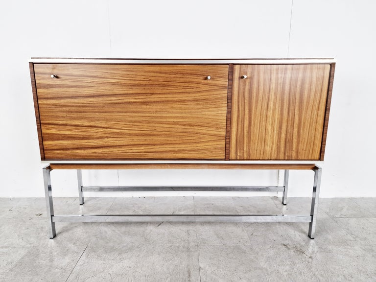 Beautiful wooden bar cabinet made in belgium in the 1960s by AL meubel and designed by Pieter De Bruyne.

It consists of a fold down door and a smaller 

Your typical mid century bar cabinet with some nice design touches.

The chrome legs are