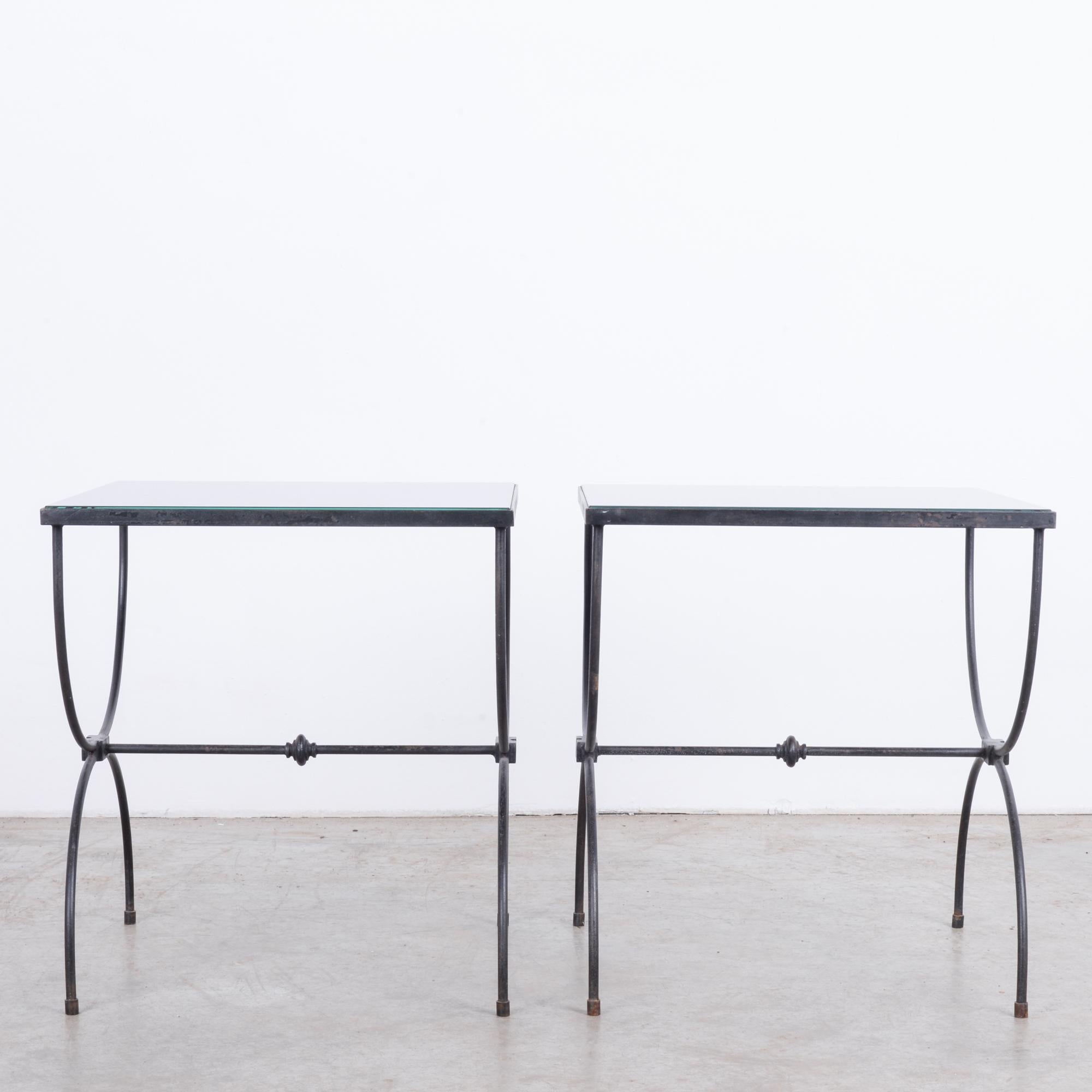 This pair of coffee tables was made in Belgium, circa 1960. Characteristic of midcentury design, the tables combine materials to feature a rectangular, opaque glass set in a metal frame. The black frames are constructed with semi-circular tubes on