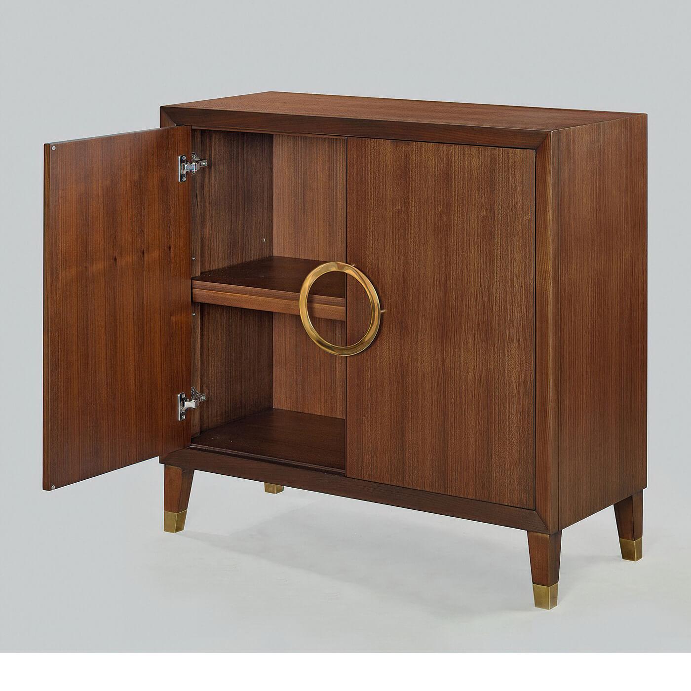 A Mid-Century Modern style walnut two-door cabinet with a beveled frame, a custom hand cast polished brass ring handle, interior shelves, raised on square tapered legs with brass caps and finished in a warm truffle stain to highlight the beautiful