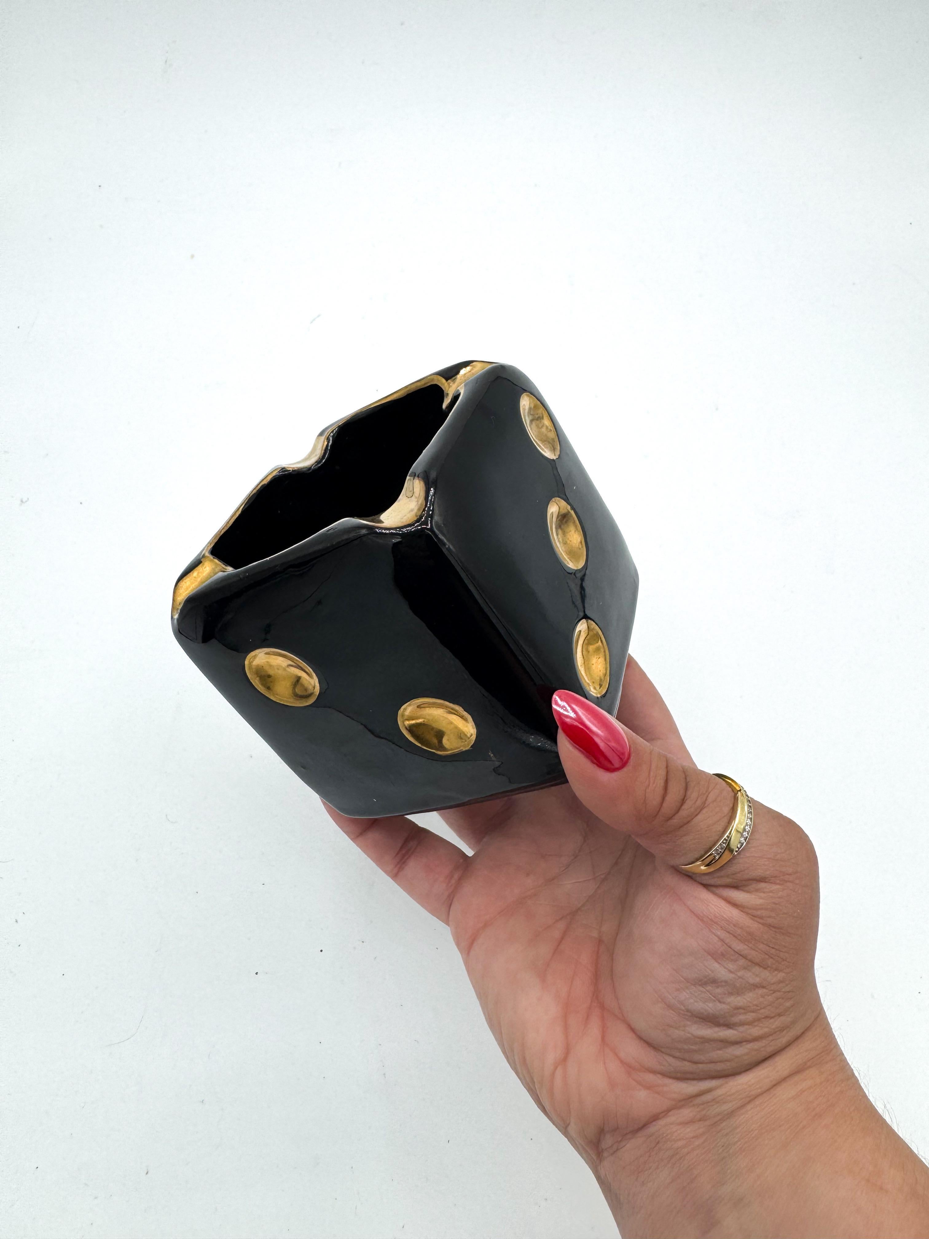 Eye-catching and whimsical, this vintage ceramic dice ashtray from the 1950s is a striking example of mid-century design. Finished in a high-gloss black glaze and accented with hand-gilded pips and cigarette rests, it merges playful form with