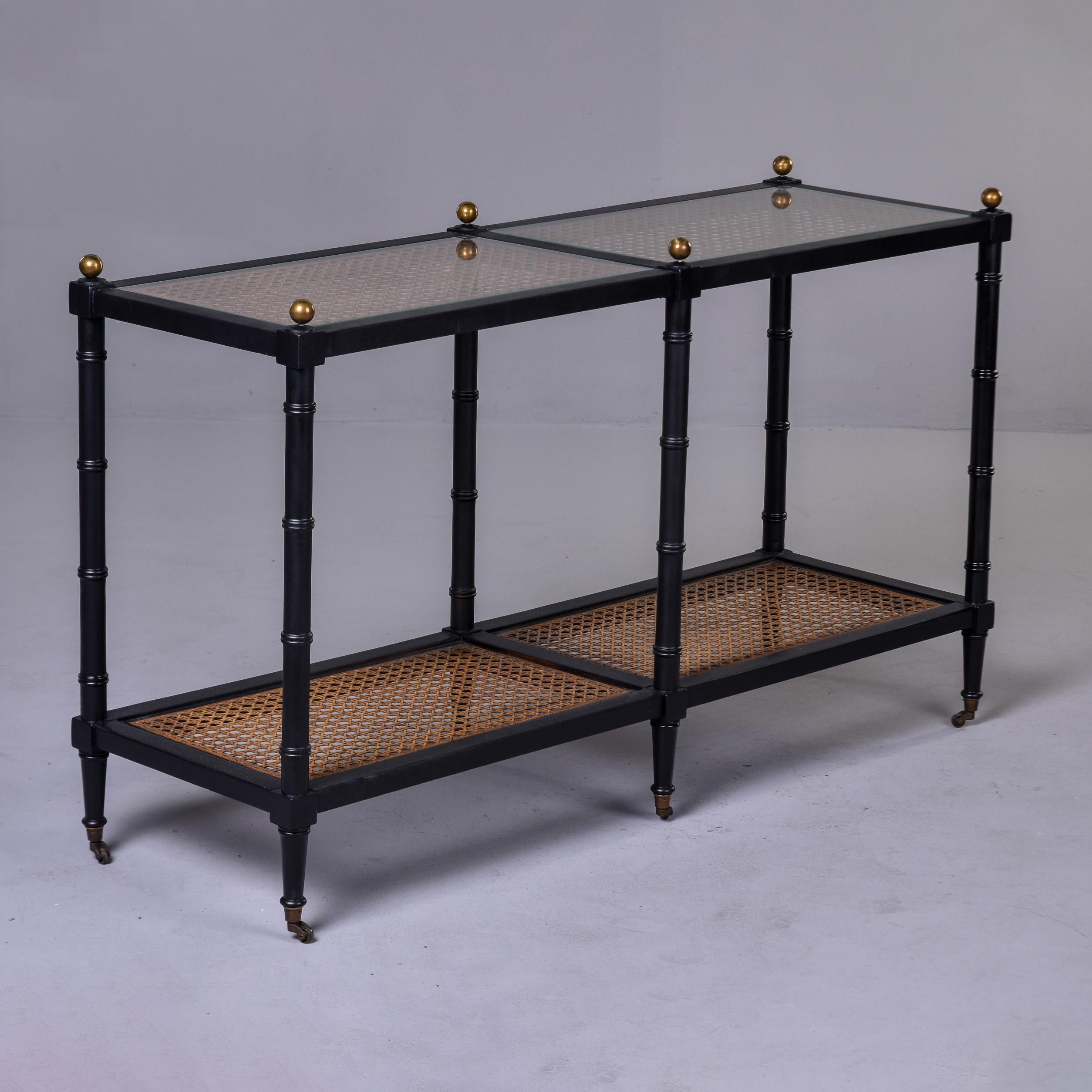 Found in the US, this circa 1960s console table has a faux bamboo frame with a painted black finish. The top and bottom shelves have caned surfaces with glass overlay panels on the top. Six legs with solid brass casters and round brass finials.