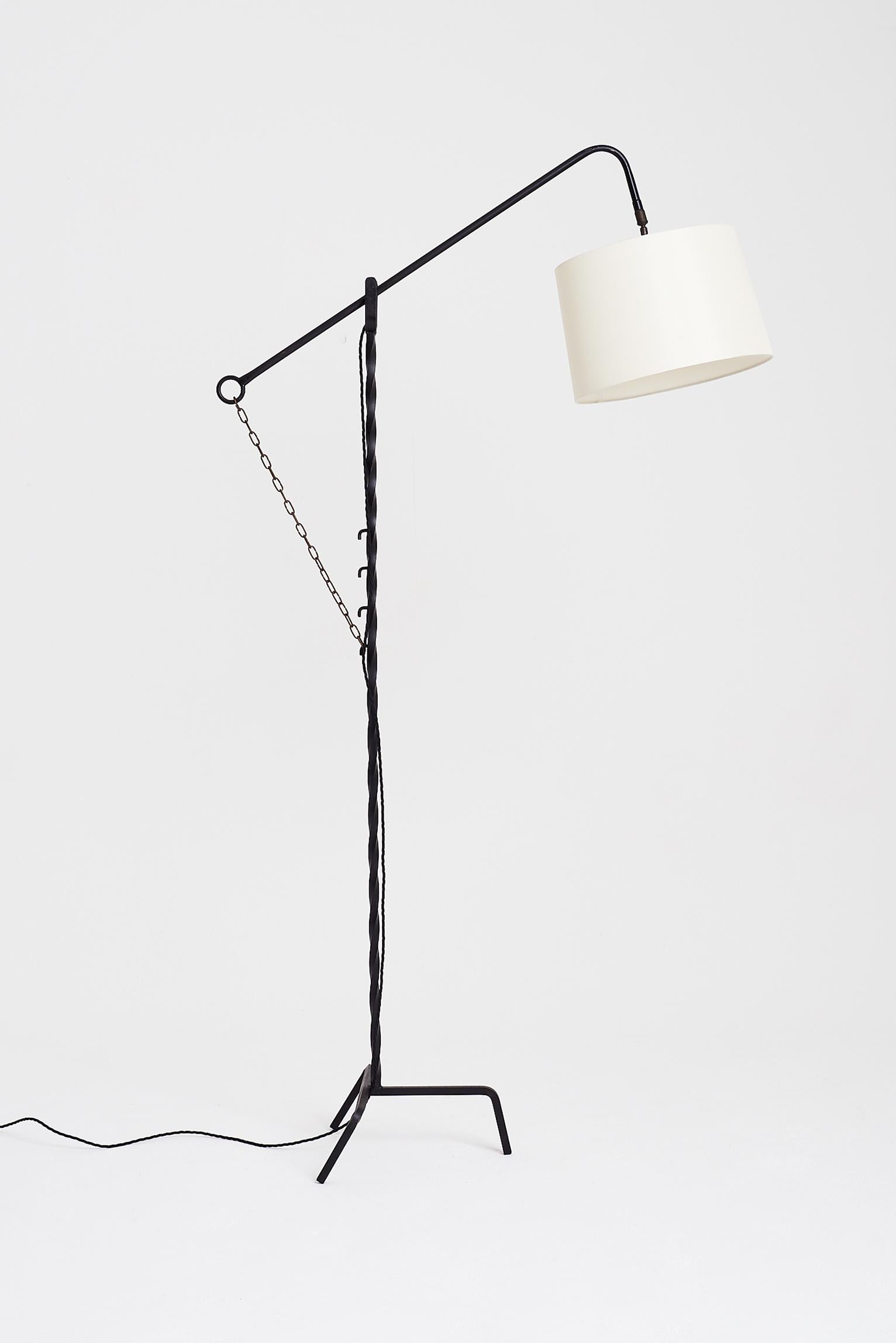 A black wrought iron floor lamp, the twisted stem on a tripod base, holding an adjustable arm hung on a cremaillere system with a brass chain.
France, circa 1950.