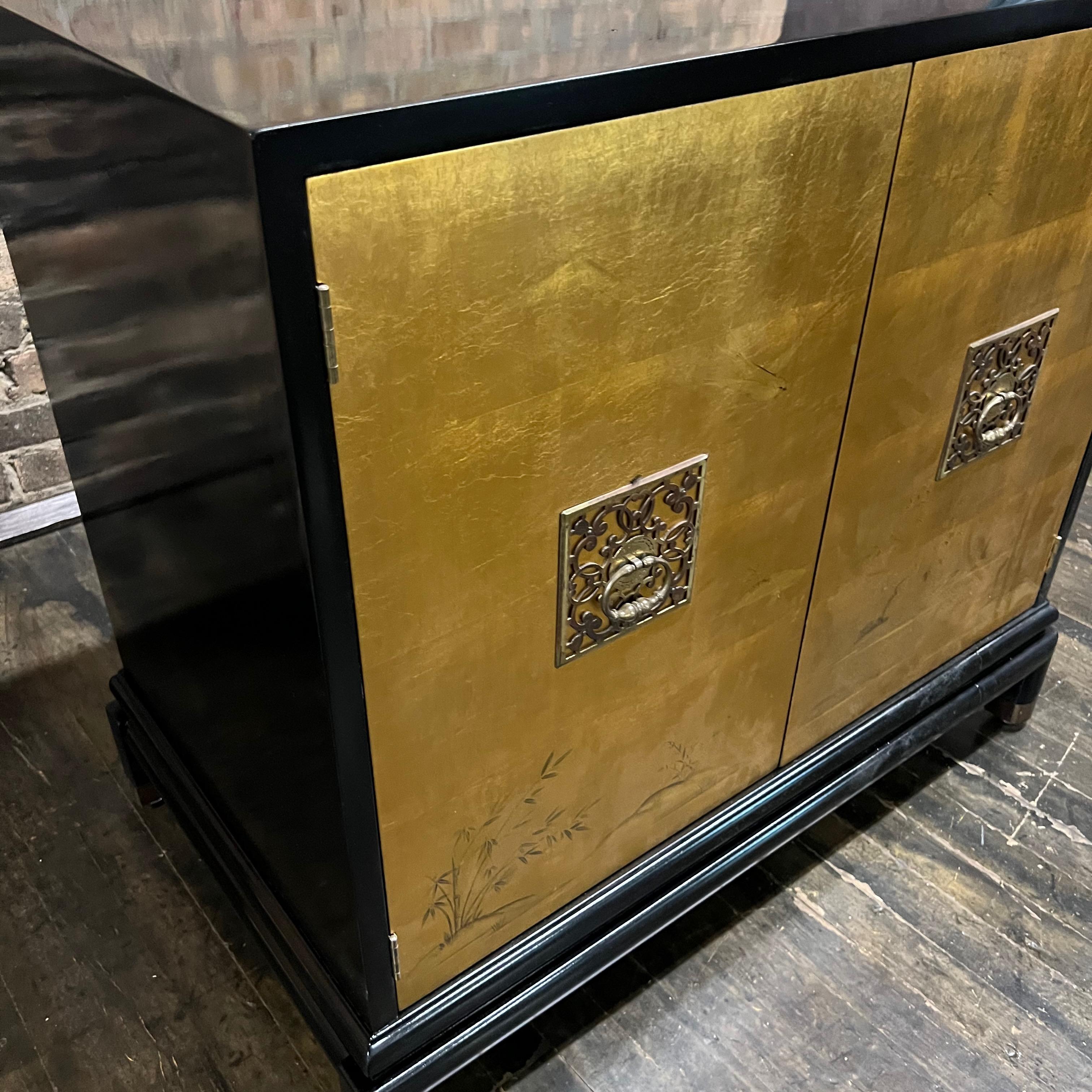 This is a stunning Chinoiserie style black lacquer cabinet with gilded (golden) doors that have images of birds, mountains and grasses painted in the gilding. There are two decorative brass door pulls. Cabinet opens to reveal one adjustable shelf.