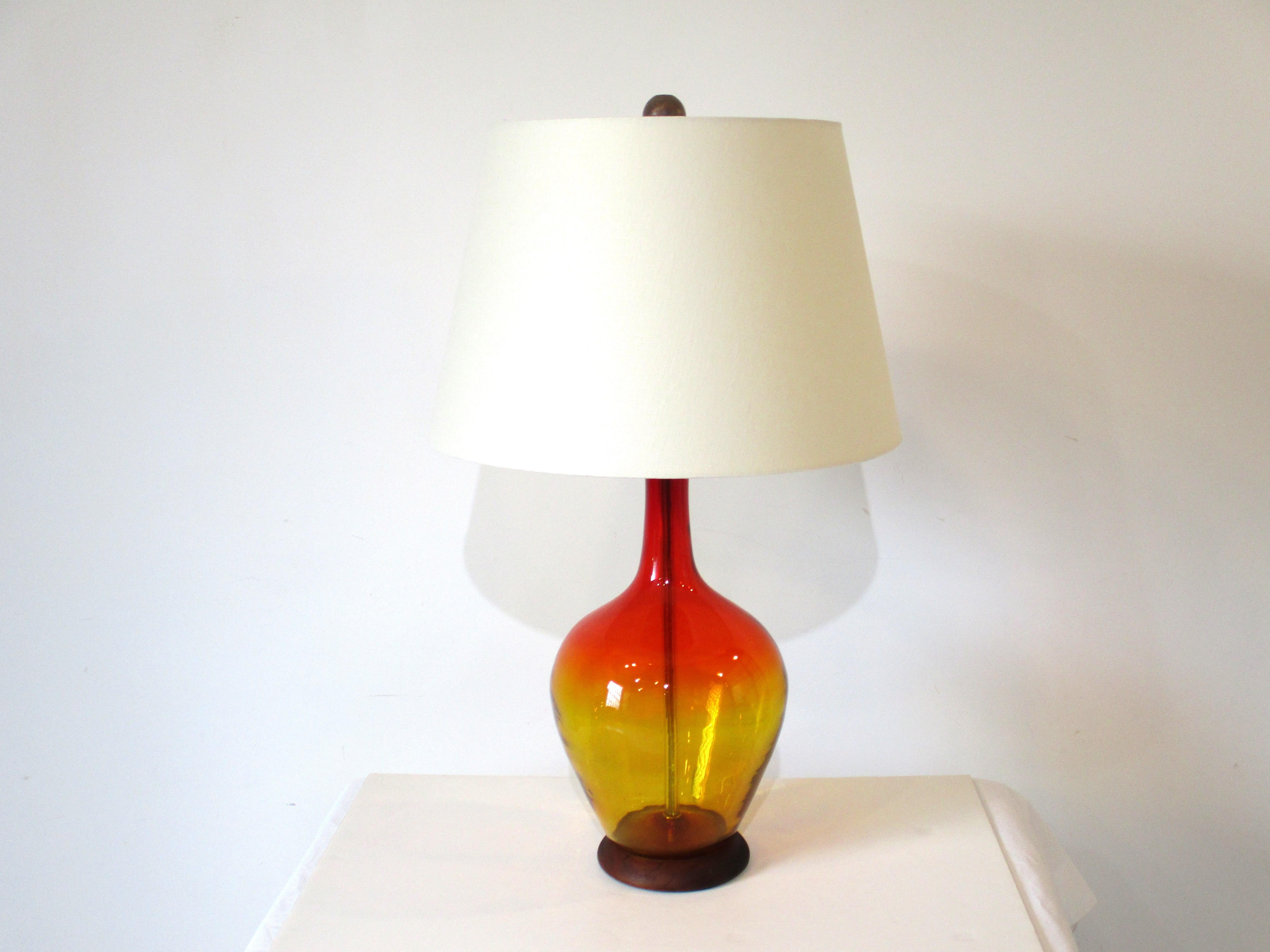 A iconic Mid Century blown glass table lamp by the Blenko glass company in a red amber fade called Amberina. Crafted by the artist Winslow Anderson and sits on a dark walnut base topped with a cream linen shade and dark wood final the finishing