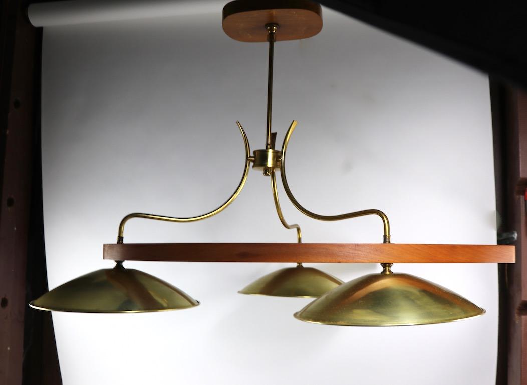 Very stylish Mid-Century Modern three-light chandelier of brass, glass and walnut. This hanging fixture has a tubular brass structure, which connects to a solid walnut boomerang, or horseshoe form body, from which hang circular brass hoods, that