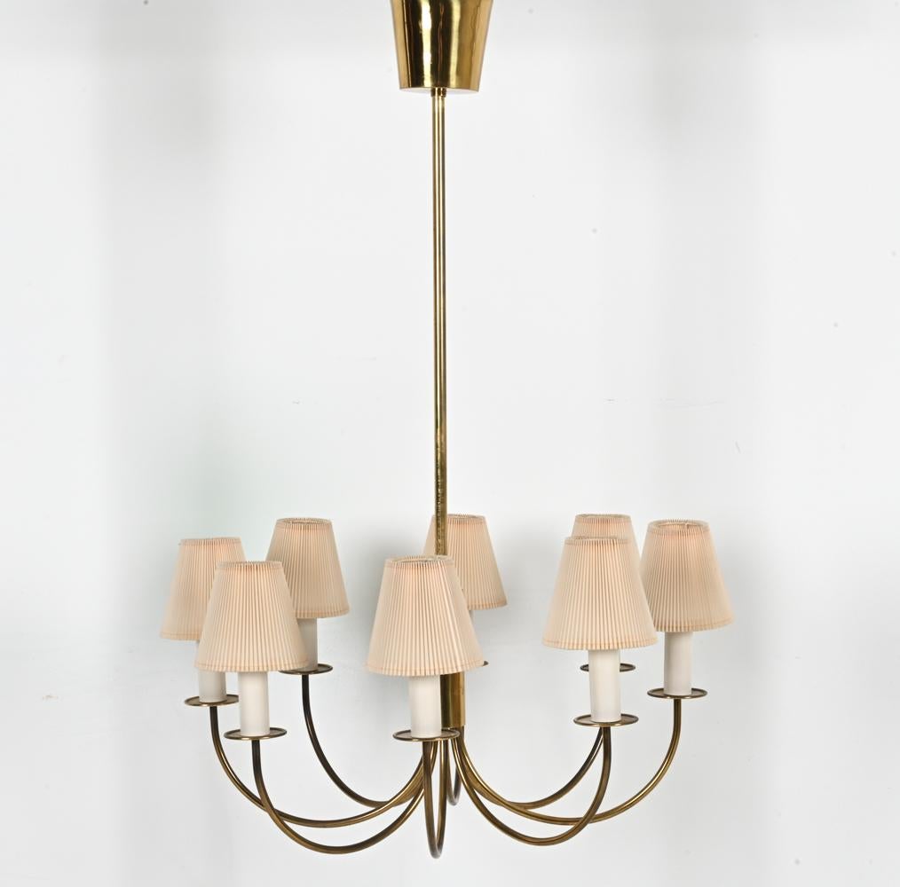 This elegant Scandinavian mid-century chandelier, attributed to Danish designer Th. Valentiner, known for his Functionalist forms, features eight U-shaped arms emanating from a central brass stem. Petite pleated shades attractively diffuse light to