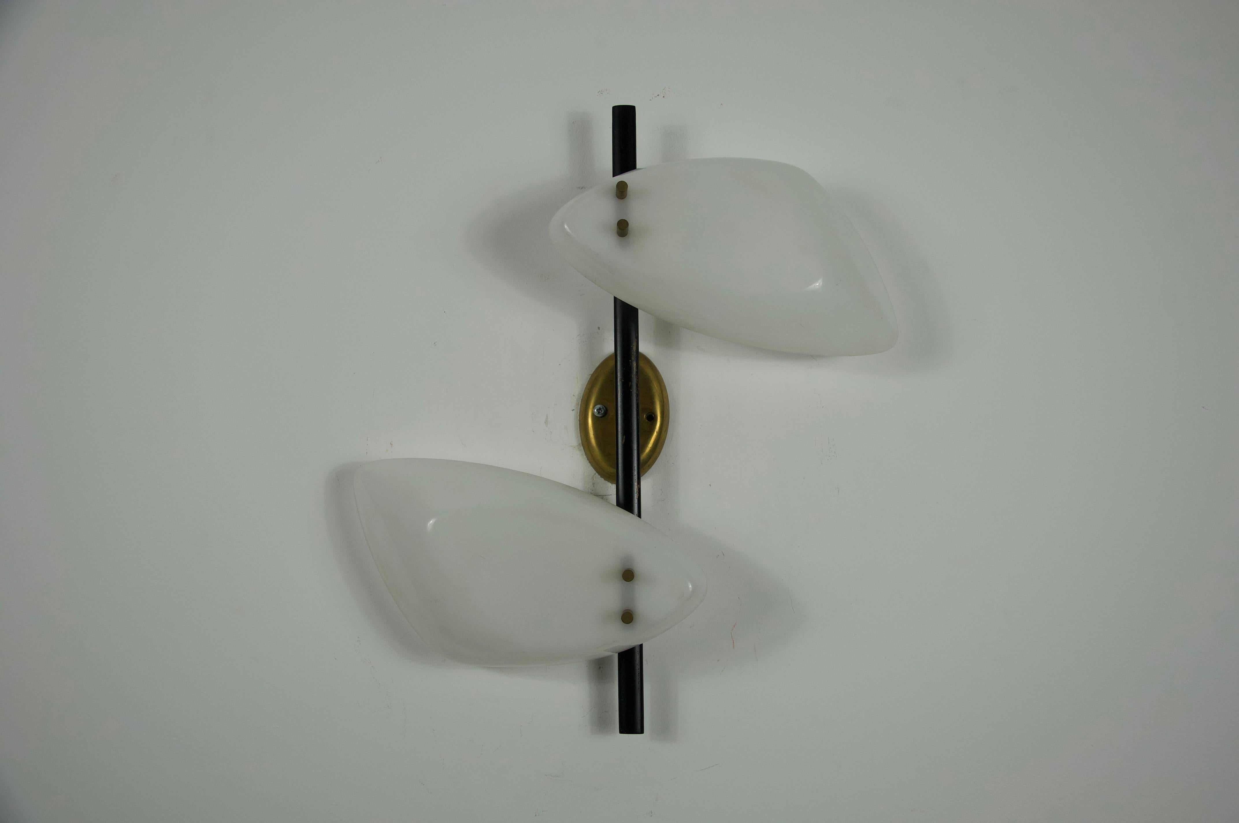 Questa sorprendente lampada da parete italiana del Mid-Century, risalente agli anni '60, mette in mostra la raffinata sensibilità del design dell'epoca. Con due paralumi asimmetrici in plexiglass bianco, montati su uno stelo in metallo nero con