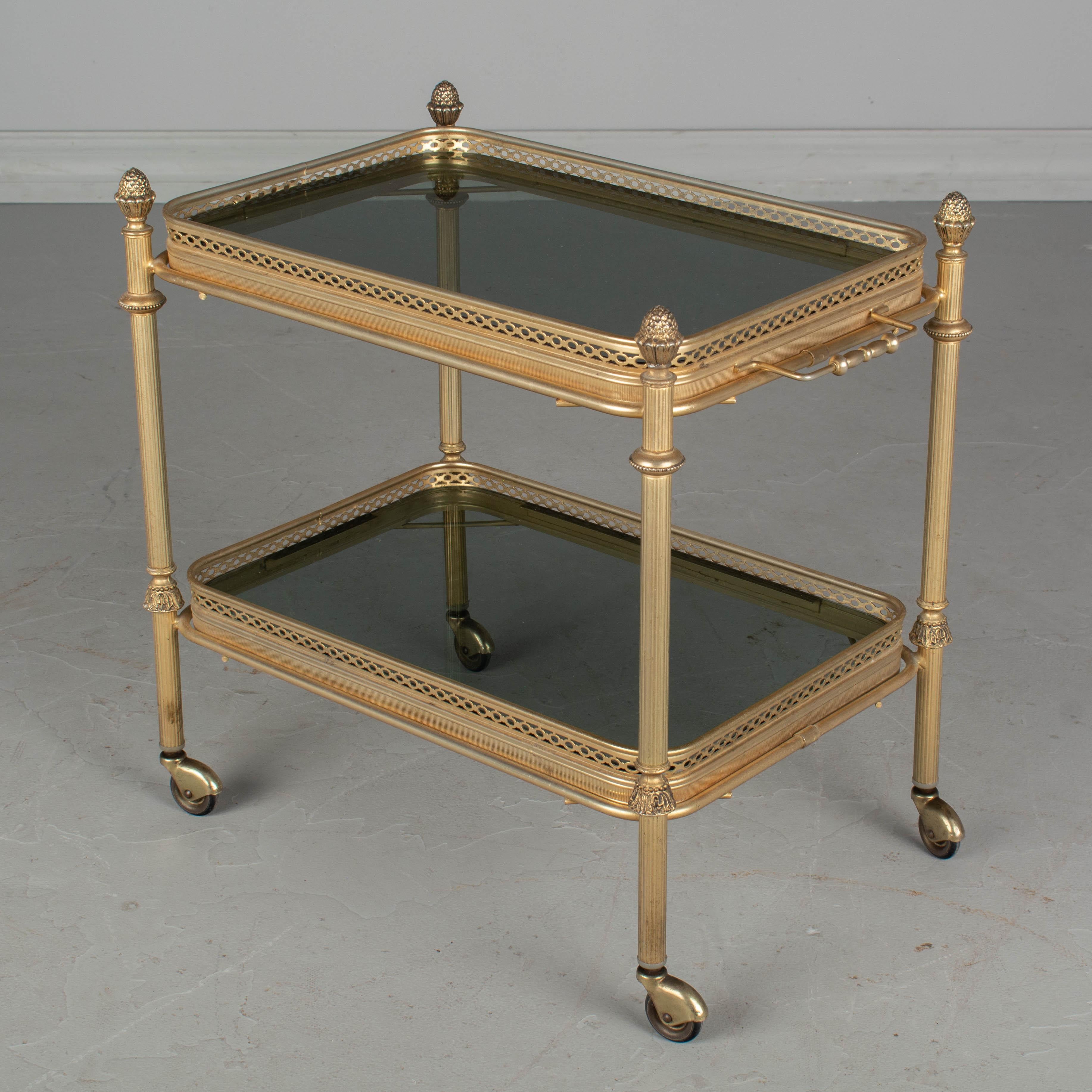 A diminutive Mid-Century Modern French bar cart, or drinks trolley, with brass plated finish and smoke glass shelves with decorative gallery. Each tray easily removes and the top tray has handles for serving drinks. Original rubber wheels glide