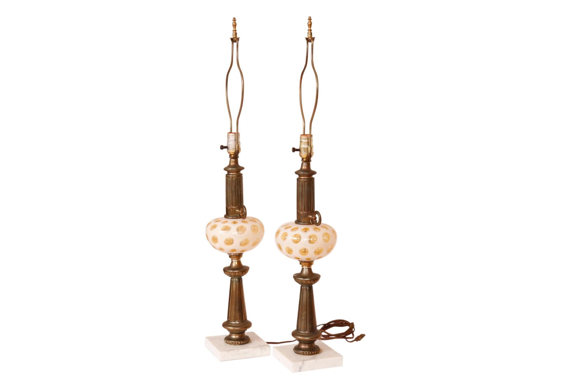 A pair of mid century brass and glass table lamps. Central brass columns are reeded with blown glass vases at the center. The glass vases are milk glass with clear yellow bubbles blown from within. Lamps stand on square marble bases. One lamp has a