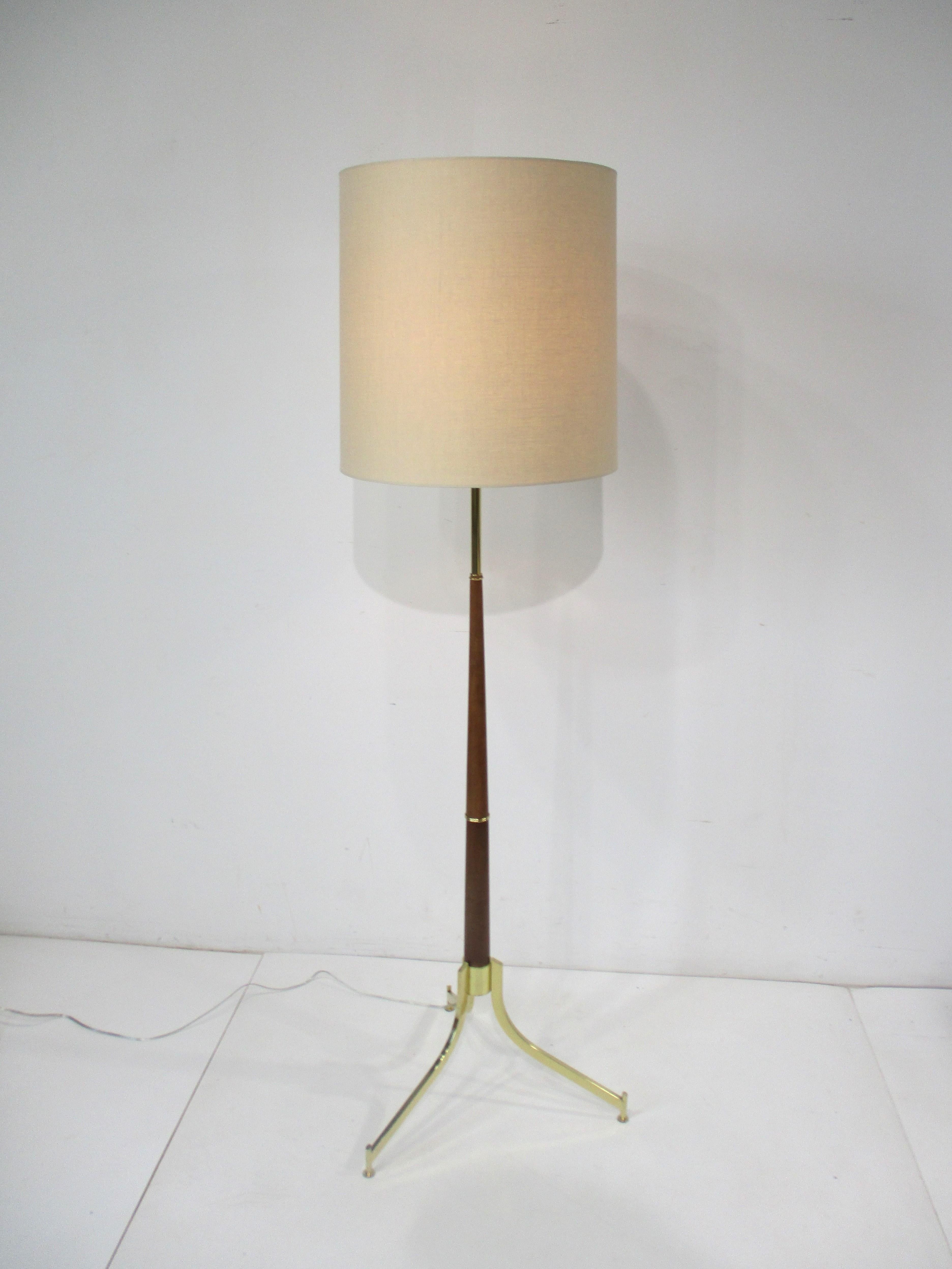 A very well designed Mid Century floor lamp with tri pod brass legs having foot pads and brass details to the center pole and upper shaft . The pole is in a rich medium toned mahogany wood and topped with a beige linen barrel lamp shade created in