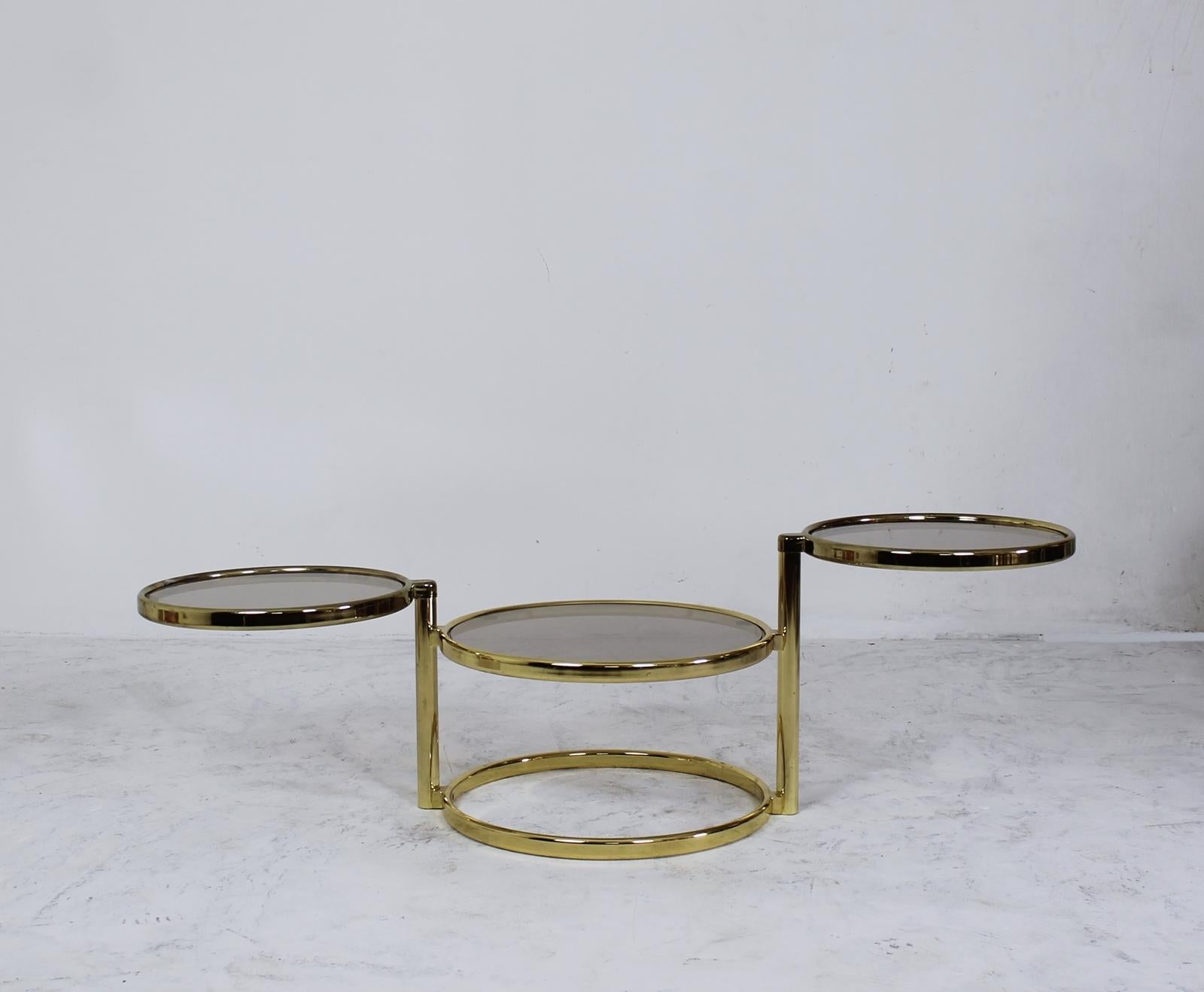 A three-tier brass plated metal structure swivel table. The top two tiers pivot to expand to a width of 132cm. Functions as a side table when closed or a coffee table when fully open. Glass has a slightly smokey tone.