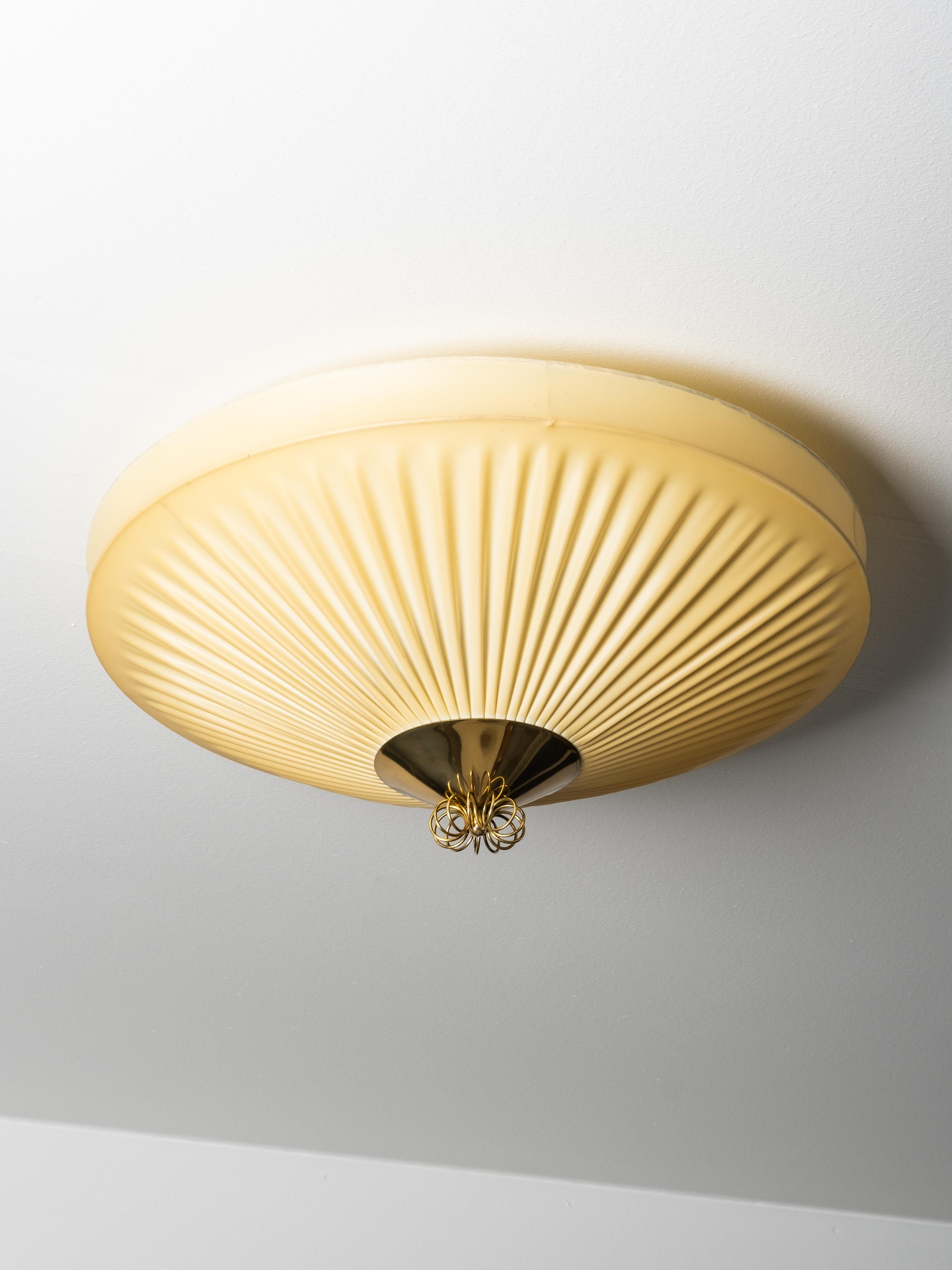 Cette applique finlandaise du milieu du siècle dernier, datant des années 1950, combine un abat-jour en textile synthétique plissé avec un épi de faîtage en laiton et un dôme central en laiton. La forme large et peu profonde crée une présence