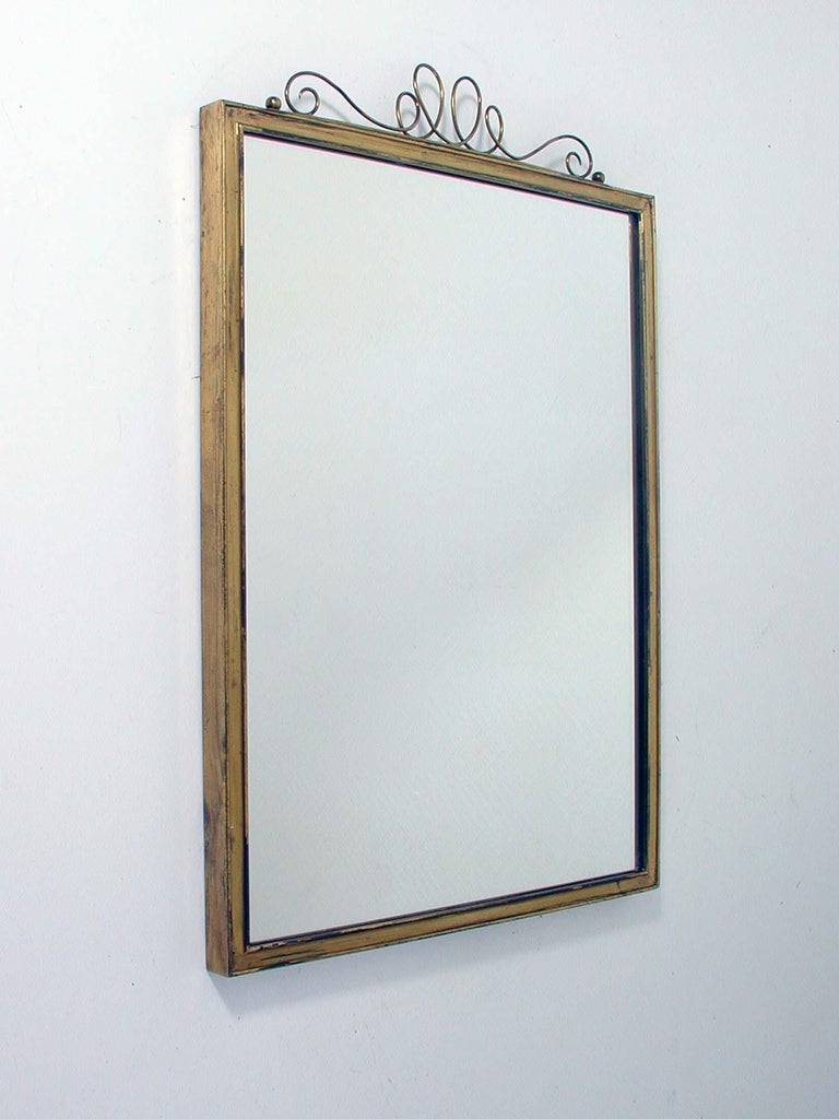 This beautiful midcentury wall mirror has got a brass frame and a looped brass finial. It was designed and manufactured in Germany in the 1950s by Münchner Zierspiegel.

The mirror is in very good condition with a really nice patina of age on the
