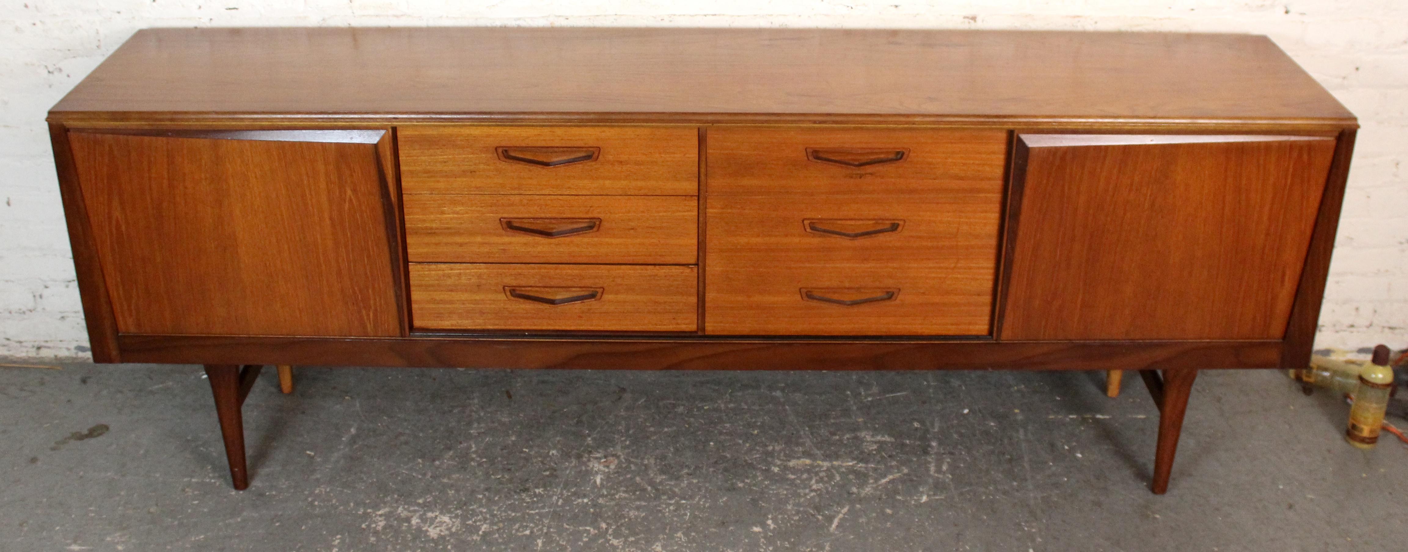 Questa credenza di Royal Heritage Furniture, realizzata in modo eccellente, è un esempio delle linee pulite e dei toni caldi del teak caratteristici del design britannico degli anni '60. Caratterizzata da una disposizione simmetrica con tre ante e
