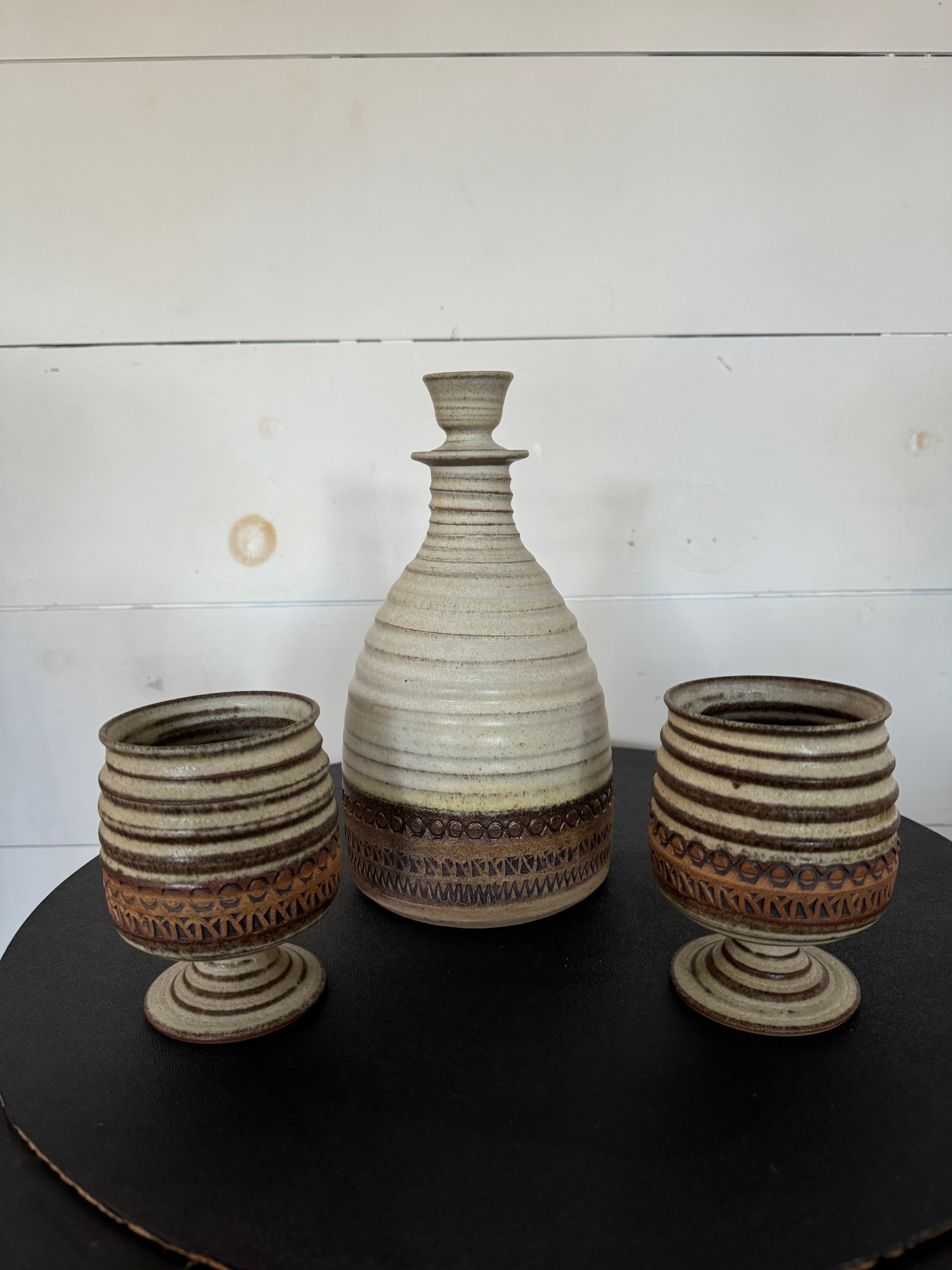 Si tratta di un set affascinante che esemplifica l'artigianato della metà del secolo scorso. Realizzato in gres, il set comprende un decanter e delle tazze abbinate, tutte caratterizzate dai caratteristici toni della terra e dalle texture tipiche