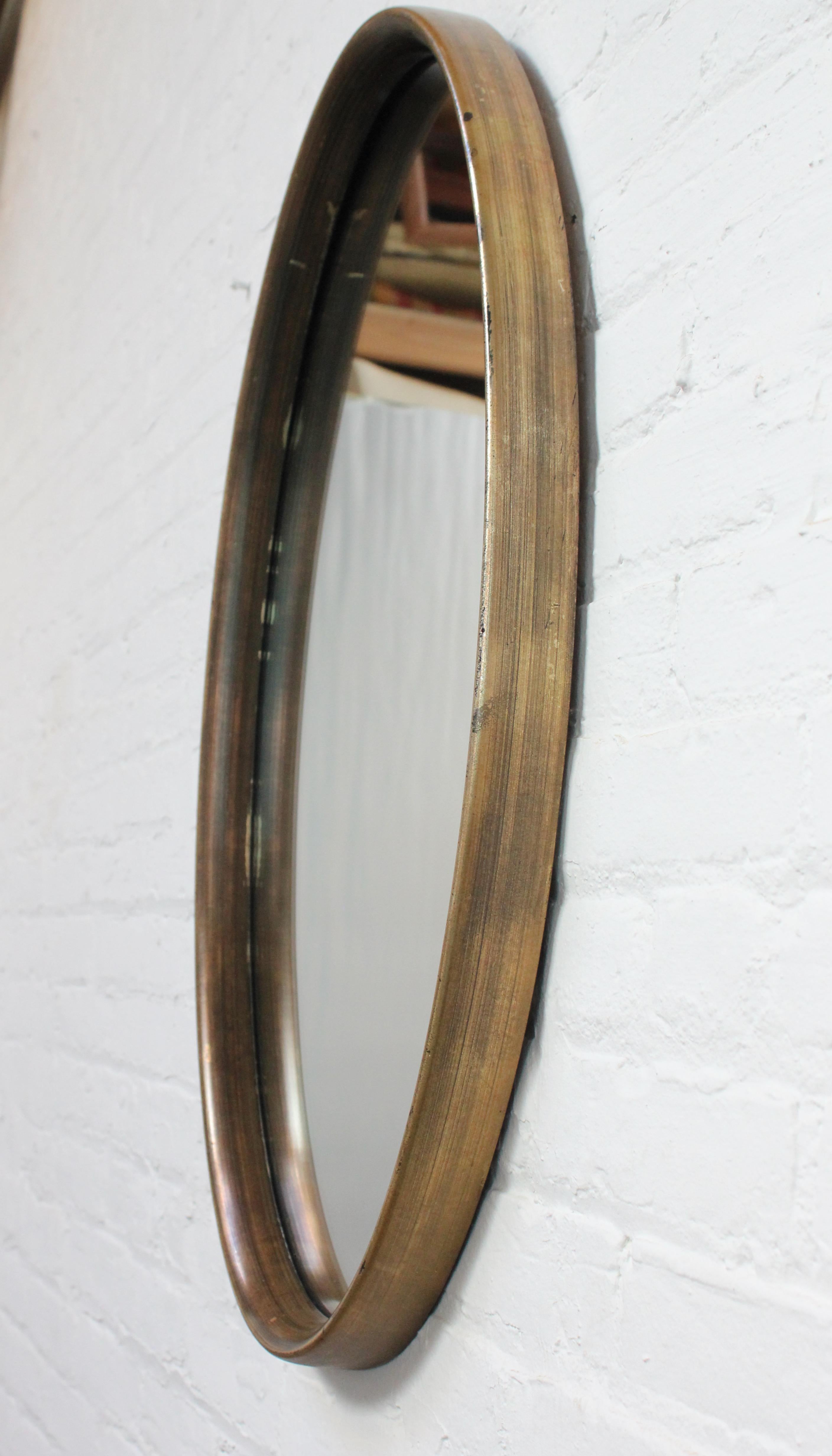 Oval wall mirror after Labarge (ca. 1970s, USA) composed of inset glass within a deep gold-painted composite frame. 
Patina throughout, producing an uneven finish (looks brown / bronze in parts and slightly more gold in others). Paint loss / age