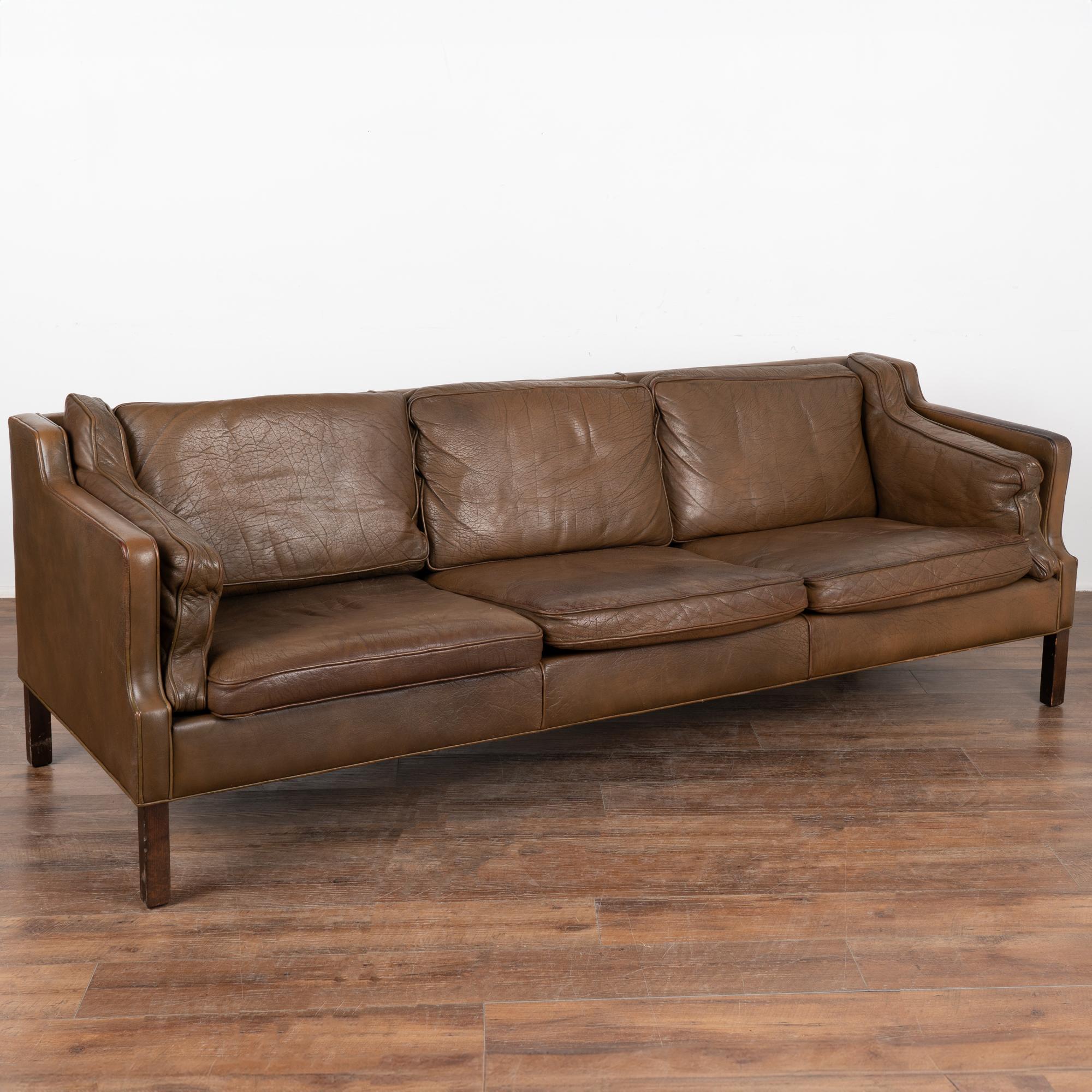 Mid-century modern brown leather three-seat sofa with removable cushions and hard wood frame.
The years of use are revealed in the aged patina of the leather, including impressions, scuffs/scratches, some discoloration, stains on cushions, multiple