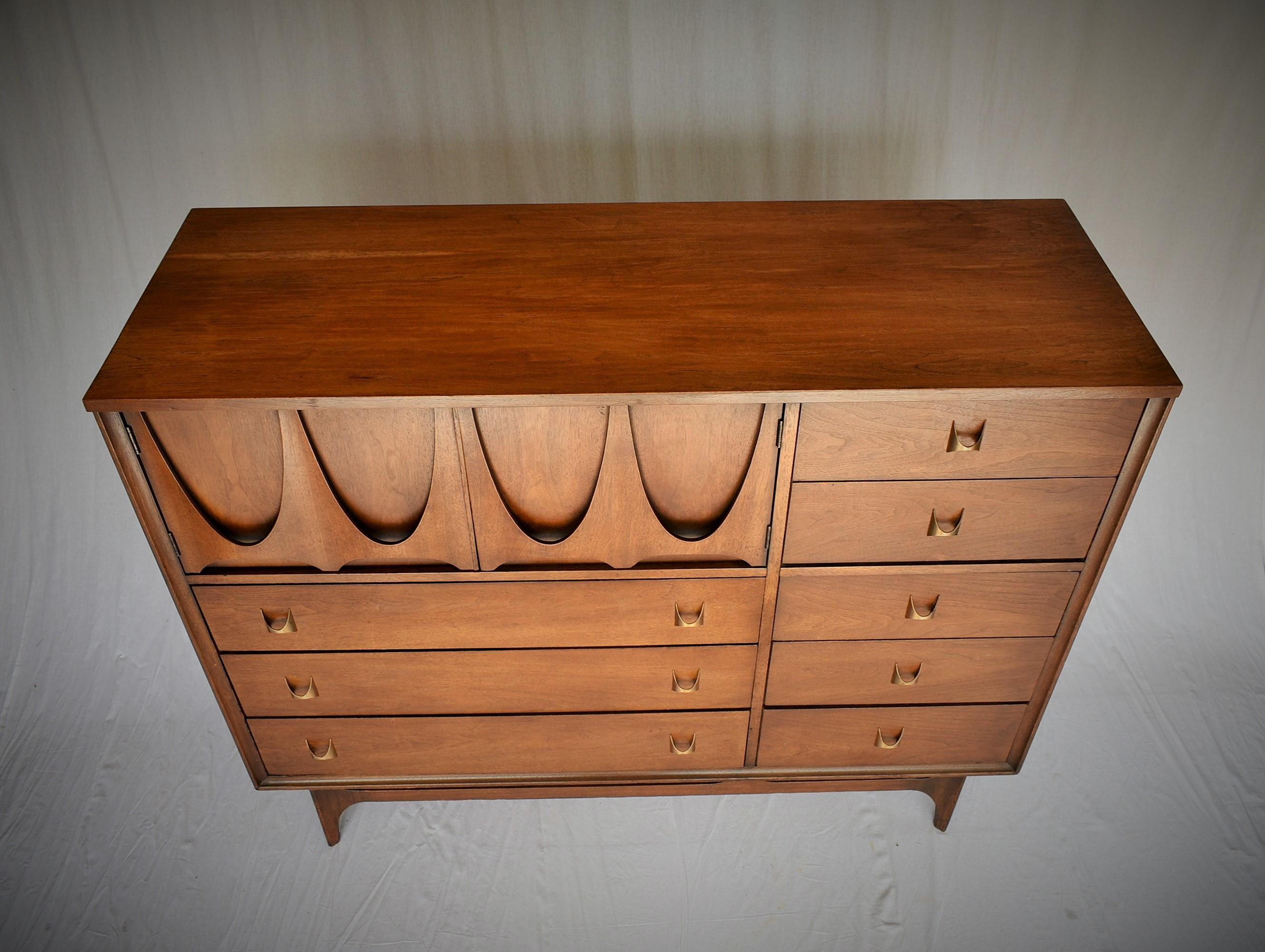 An outstanding mid-century walnut highboy dresser or chest of drawers from the Broyhill Brasilia line, circa 1960s. The chest features eight dovetailed drawers with signature brass handles. Includes open storage with two dividers behind sculpted