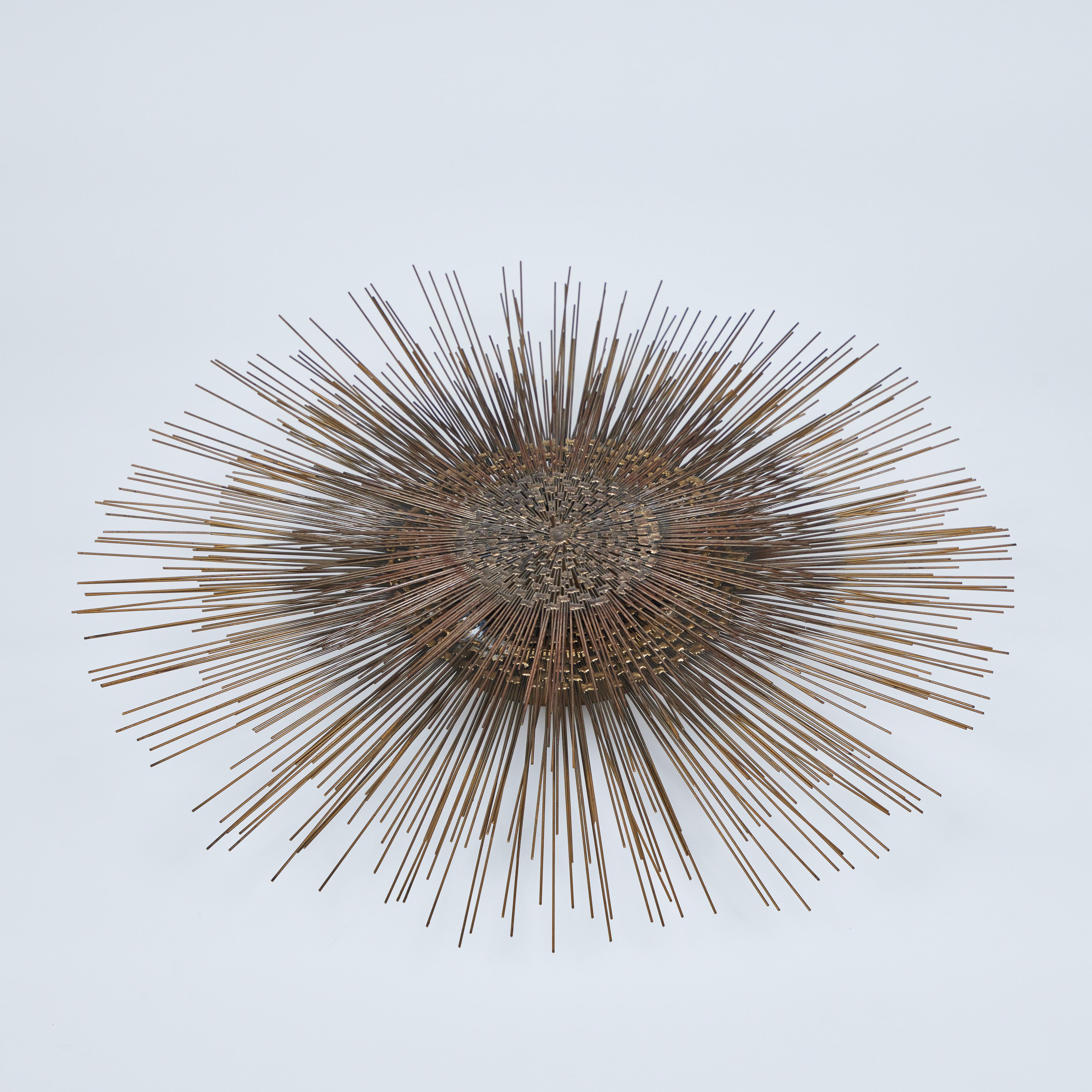 This mid century Brutalist 2-tier metal starburst wall sculpture is a great accent for any wall in any room! It is made up of 2 layers of a graduated size flat woven centers with unfinished metal rods spraying outward creating this large spacial