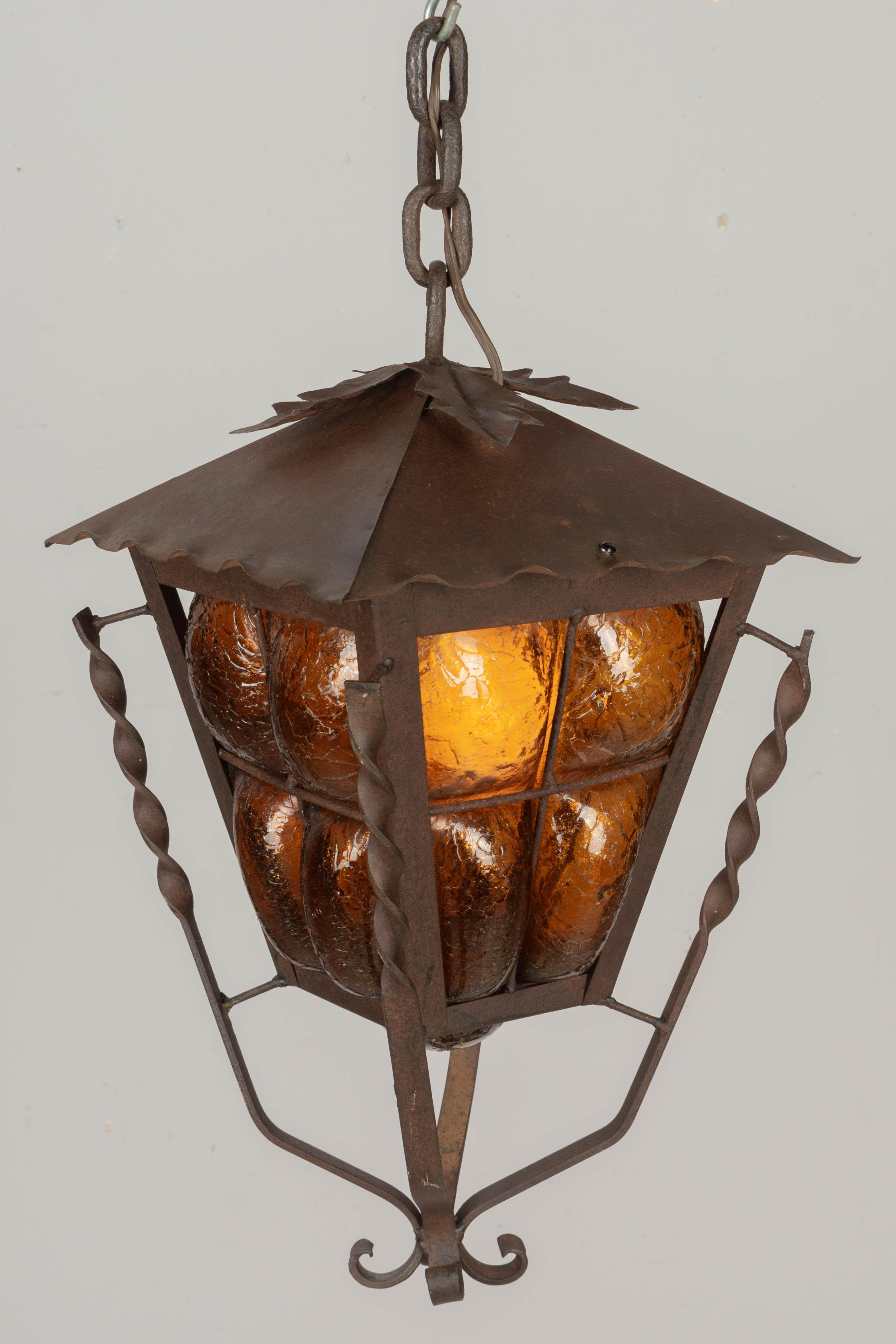 A Mid Century Mexican Arts and Crafts style caged glass hanging lantern pendant light with hand forged iron and tole frame. Thick hand-blown amber crackle glass. Tôle leaf decoration on top. Wired and in working condition. Beautiful rusted patina.