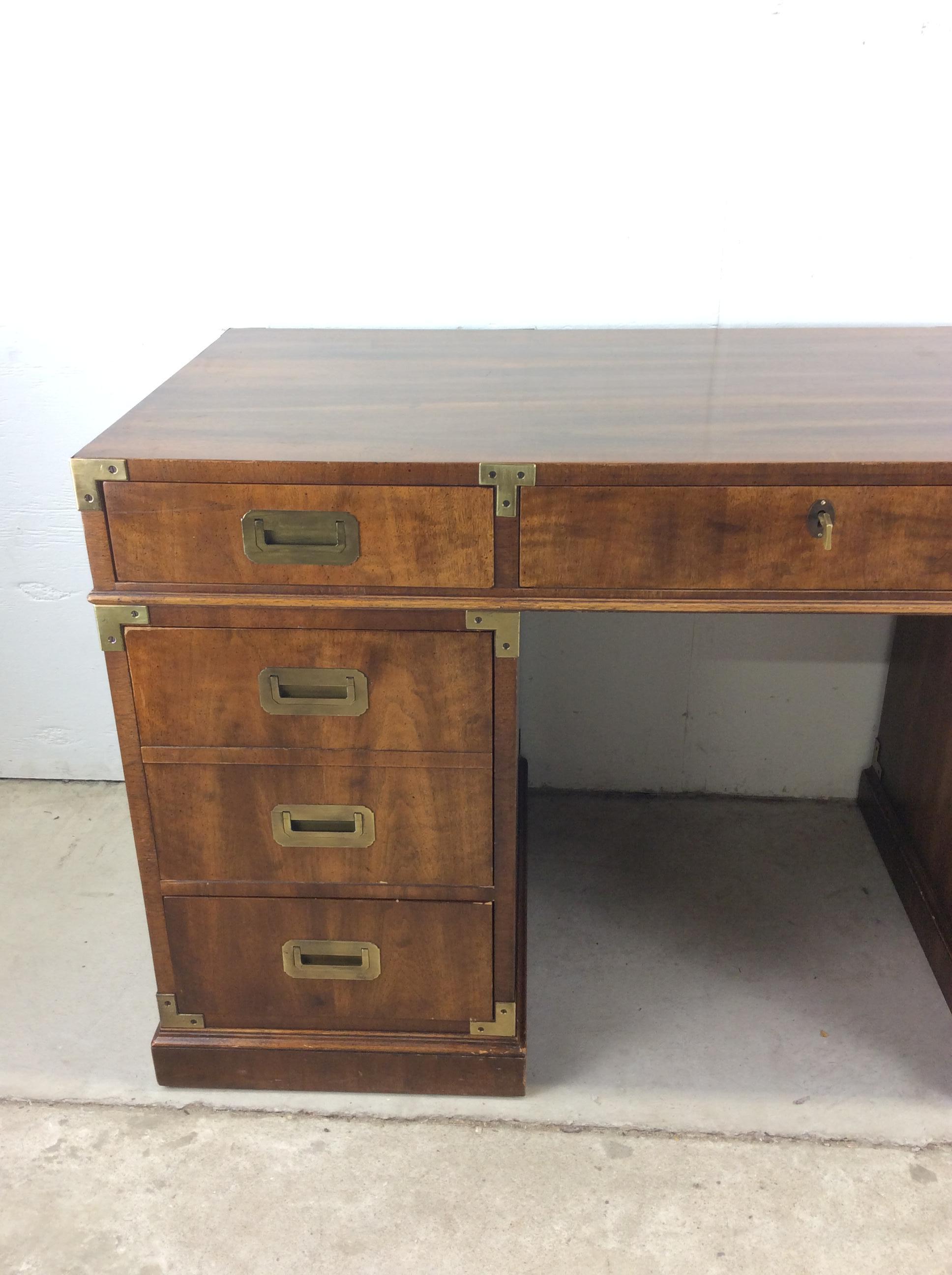 This mid century campaign style executive desk by Henredon features hardwood construction, walnut veneer with original finish, 7 dovetailed drawers with brass hardware, brass accents on the corners, locking top drawer with key included, and finished