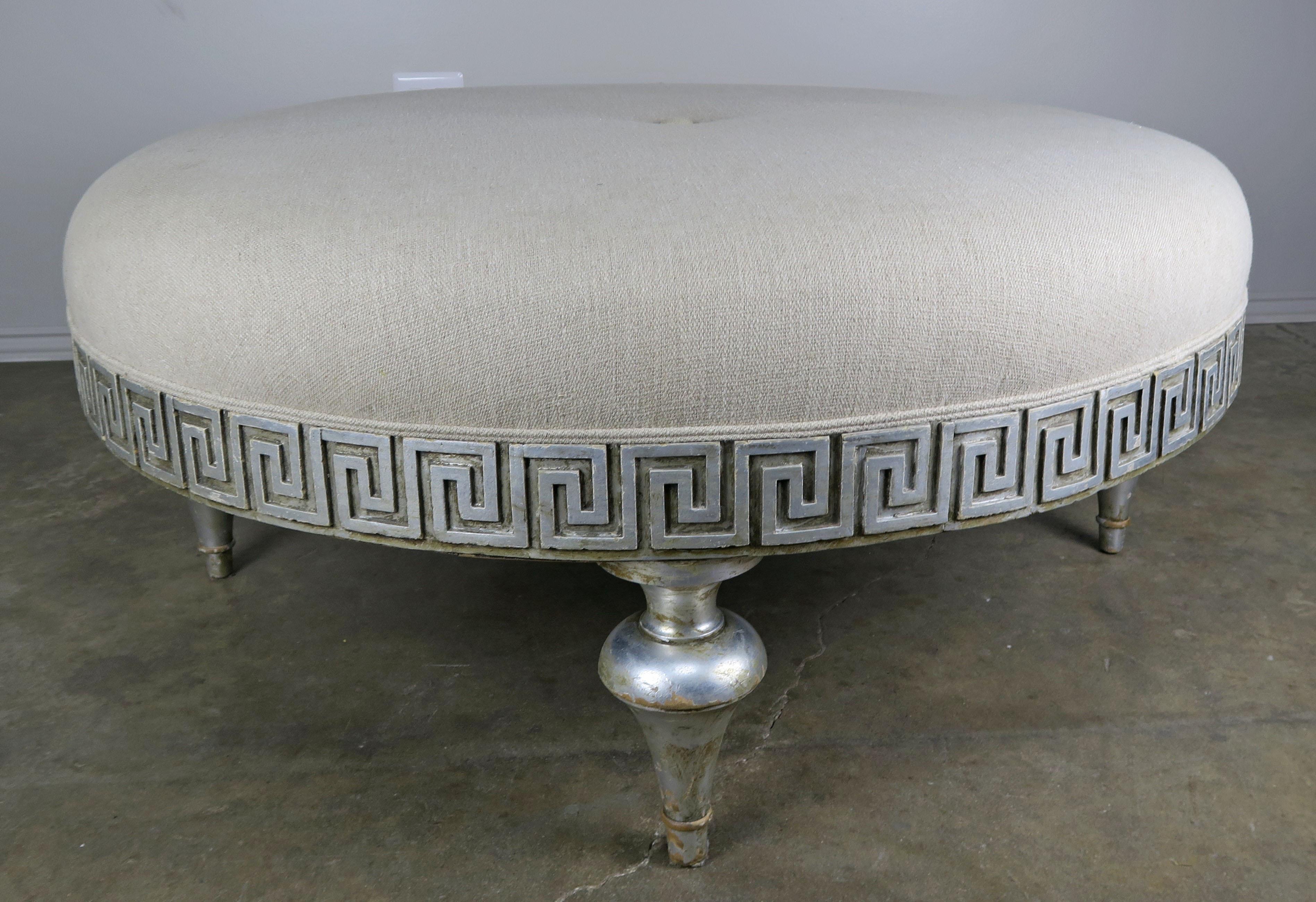 Midcentury carved silver leaf Greek key round shaped linen upholstered ottoman with self cording and center button detail. The ottoman stands on four legs.