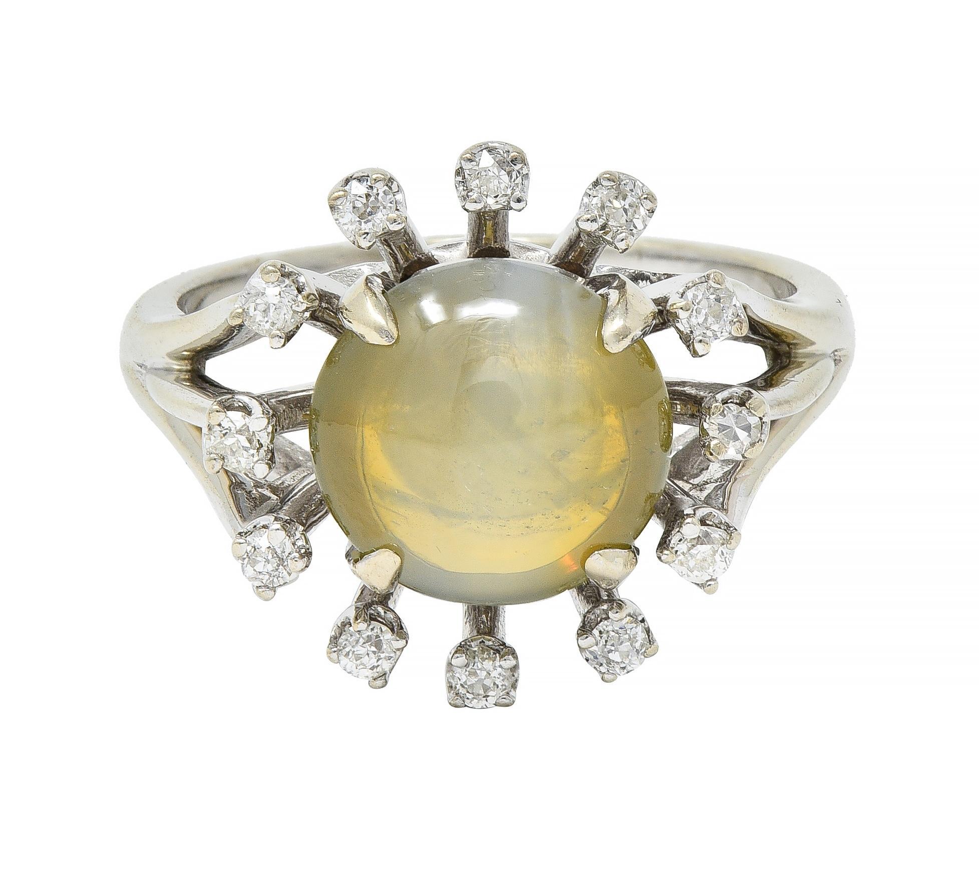 Centering a prong set round shaped cat's eye chrysoberyl cabochon measuring 8.5 mm round
Translucent yellowish green in color displaying linear chatoyancy with milk and honey effect
With a floating halo surround of prong set single cut