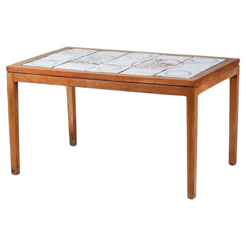 French mid-century coffee table featuring a tiled surface set within a clean, understated oak frame. The top is composed of hand-painted ceramic tiles, each etched with abstract and animal-like motifs in tones of red, blue, green, and black. The