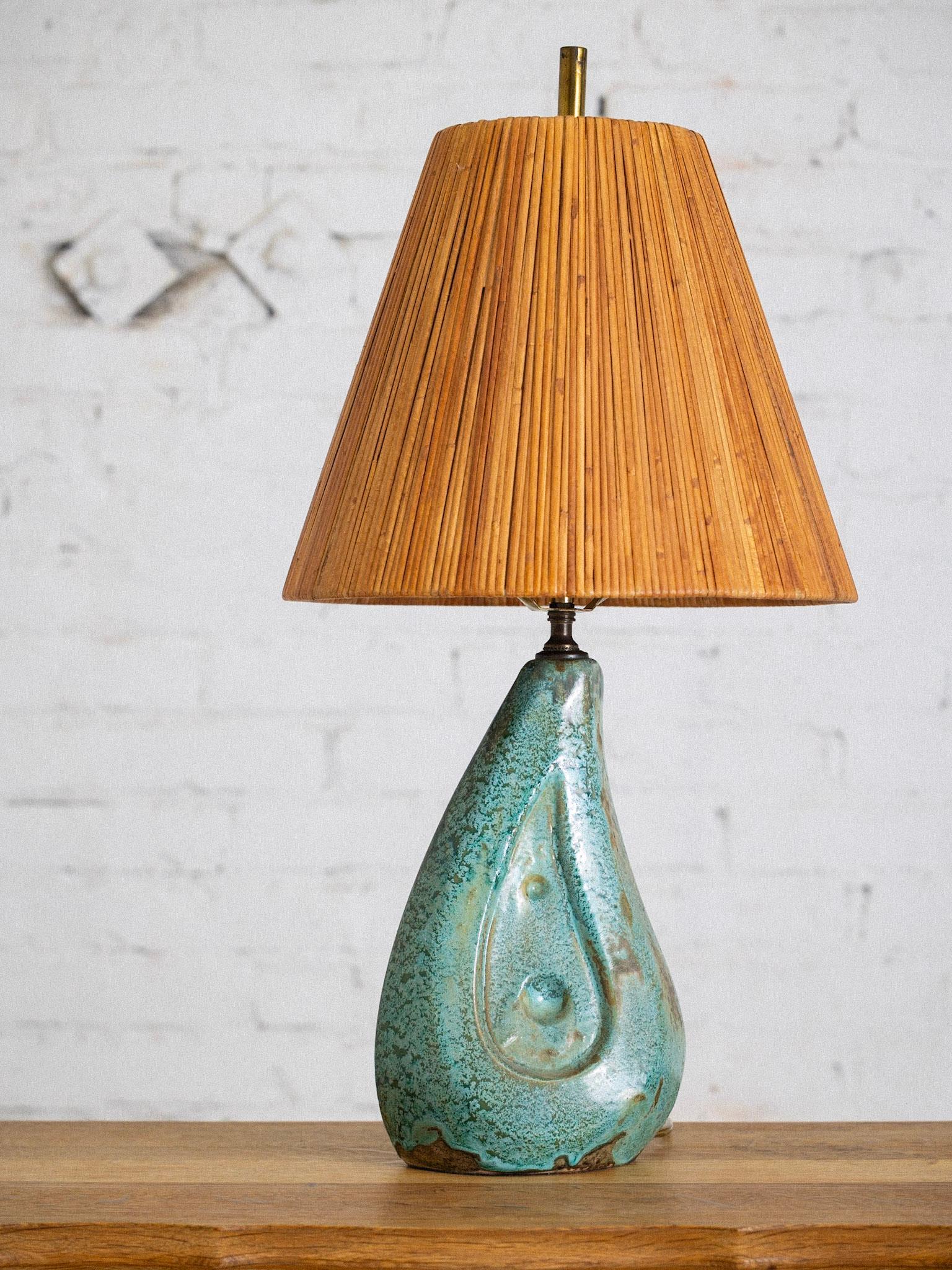 A mid century ceramic lamp with rattan wrapped shade. Tear drop form with a textured teal glaze.

Base measures 8” wide, 5” deep, 15.5” high.