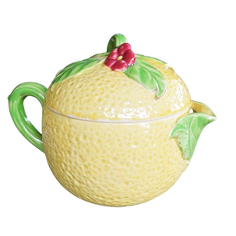 A rare ceramic juicing pitcher in the shape of a lemon. Glazed in yellow, the exterior of this pitcher is textured to mimic the skin of a lemon. The lid is round and is topped with bright green leaves, white flowers, and red berries. When removed,
