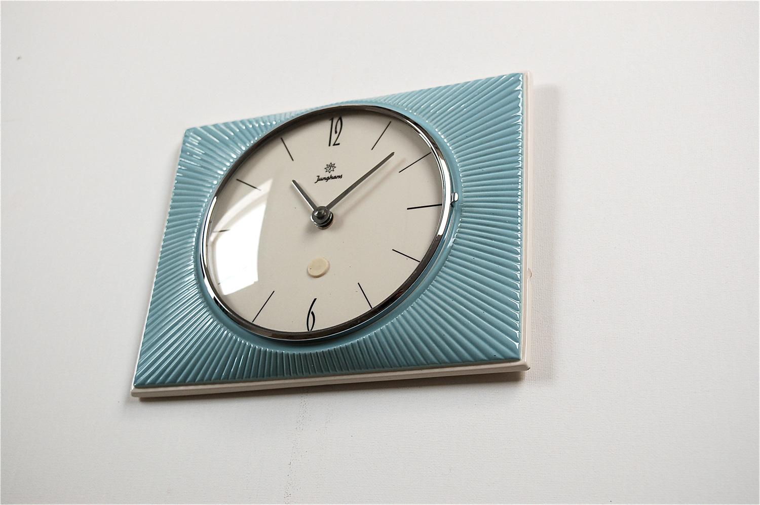 1960s, vintage ceramic wall clock by German manufacturer Junghans. The ceramic base is cream colored with a baby blue glazed front decorated in a raised sunburst pattern with the clock at the centre. It has a hinged glass front with a thin chrome