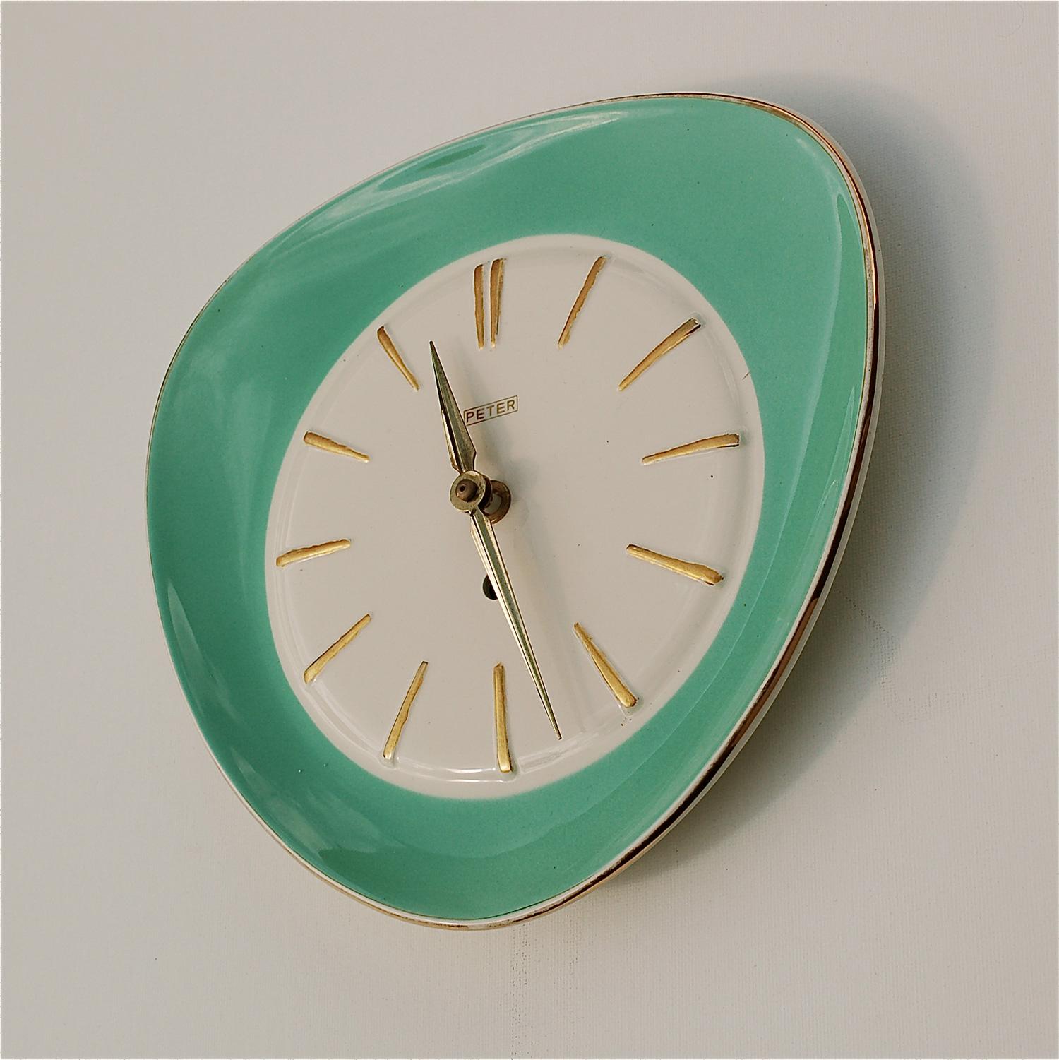 1960s vintage ceramic wall clock with a turquoise glazed front and gold trim. It is triangular in shape with curved corners and has an open clock face. The makers name Peter is featured at the top of the dial. The slightly raised stripes round the