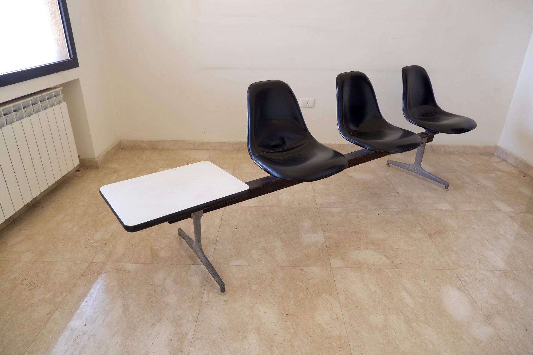 Iconic three-shell seating tandem by Charles & Ray Eames, black fiberglass shells on an aluminium base with a white side table attached, labeled on bottoms.