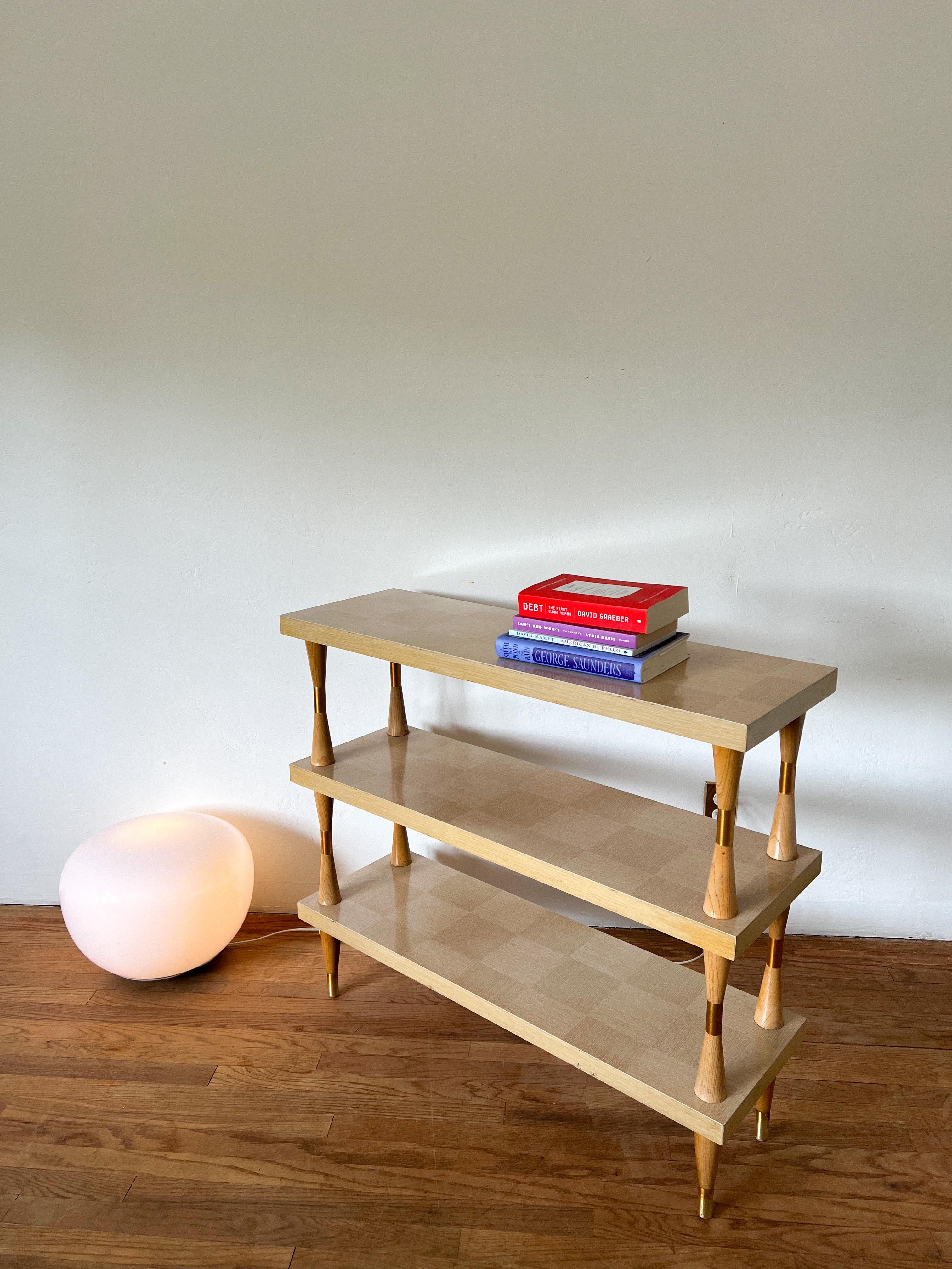 Audace scaffale in legno a tre livelli con un sorprendente motivo di impiallacciatura a scacchiera e scultoree gambe a fuso con accenti in ottone. Probabilmente risalente agli anni '60-'70, questo pezzo fonde l'artigianato moderno della metà del