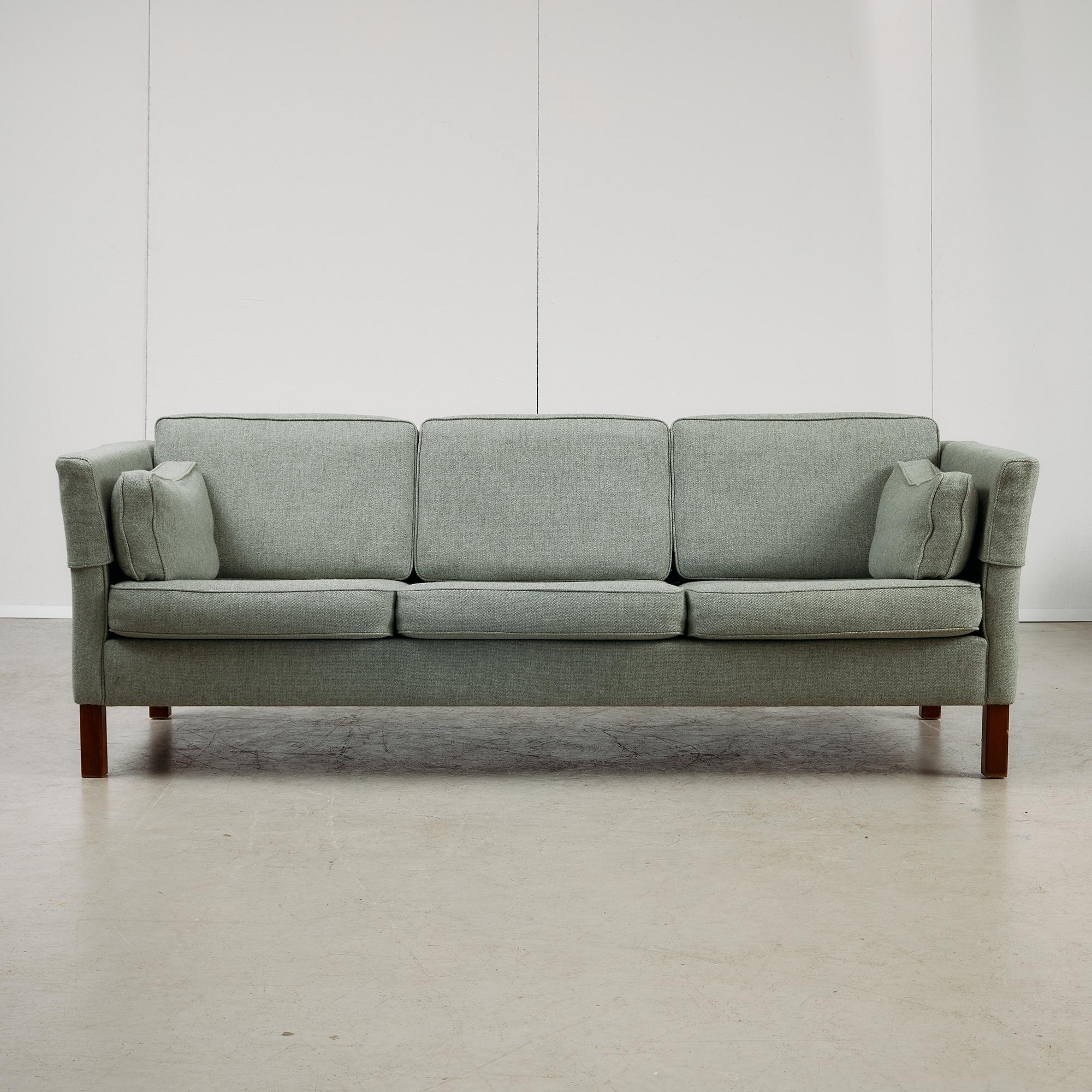 Questo divano a tre posti della metà del secolo scorso abbina linee pulite a un'attenta funzionalità. Sostenuta da gambe in legno di ciliegio, la struttura è rivestita in un tessuto grigio-verde che offre una presenza morbida e neutra. Un ulteriore