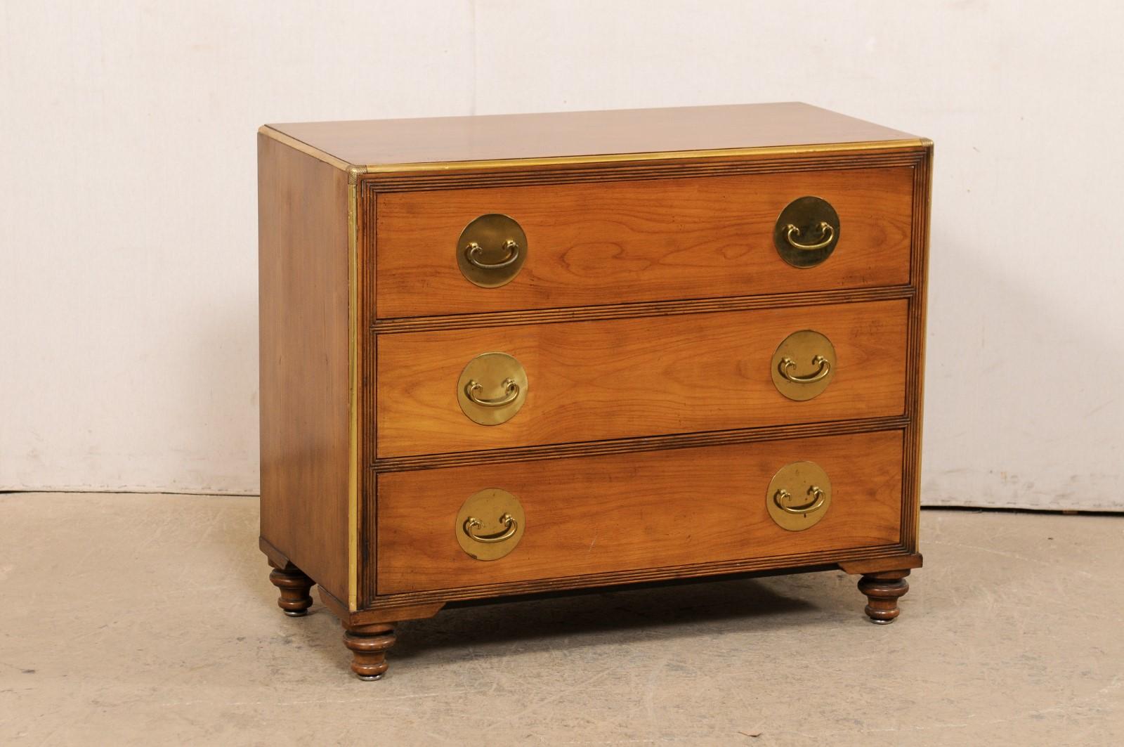 An American mid-century chest of three-drawers with brass accents. This mid 20th century chest has a rectangular-shaped top with edging along front and two sides being trimmed in brass, atop a case which houses three full-sized graduated and