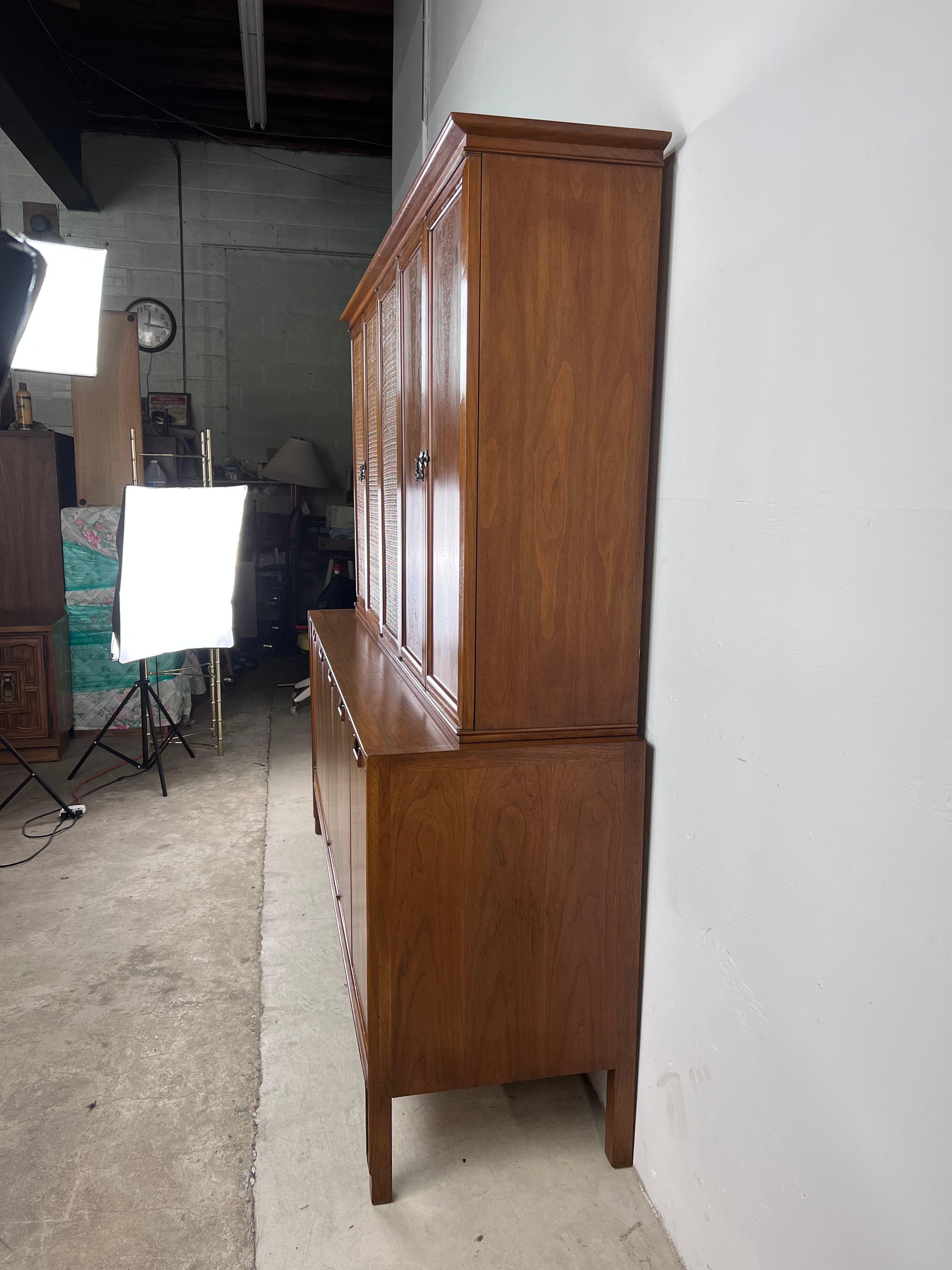 Ce vaisselier moderne du milieu du siècle de la collection Janus de Mt. Airy Furniture présente une construction en bois dur, un placage en noyer avec un fini original, un design en deux parties, trois tiroirs à queue d'aronde avec un compartiment