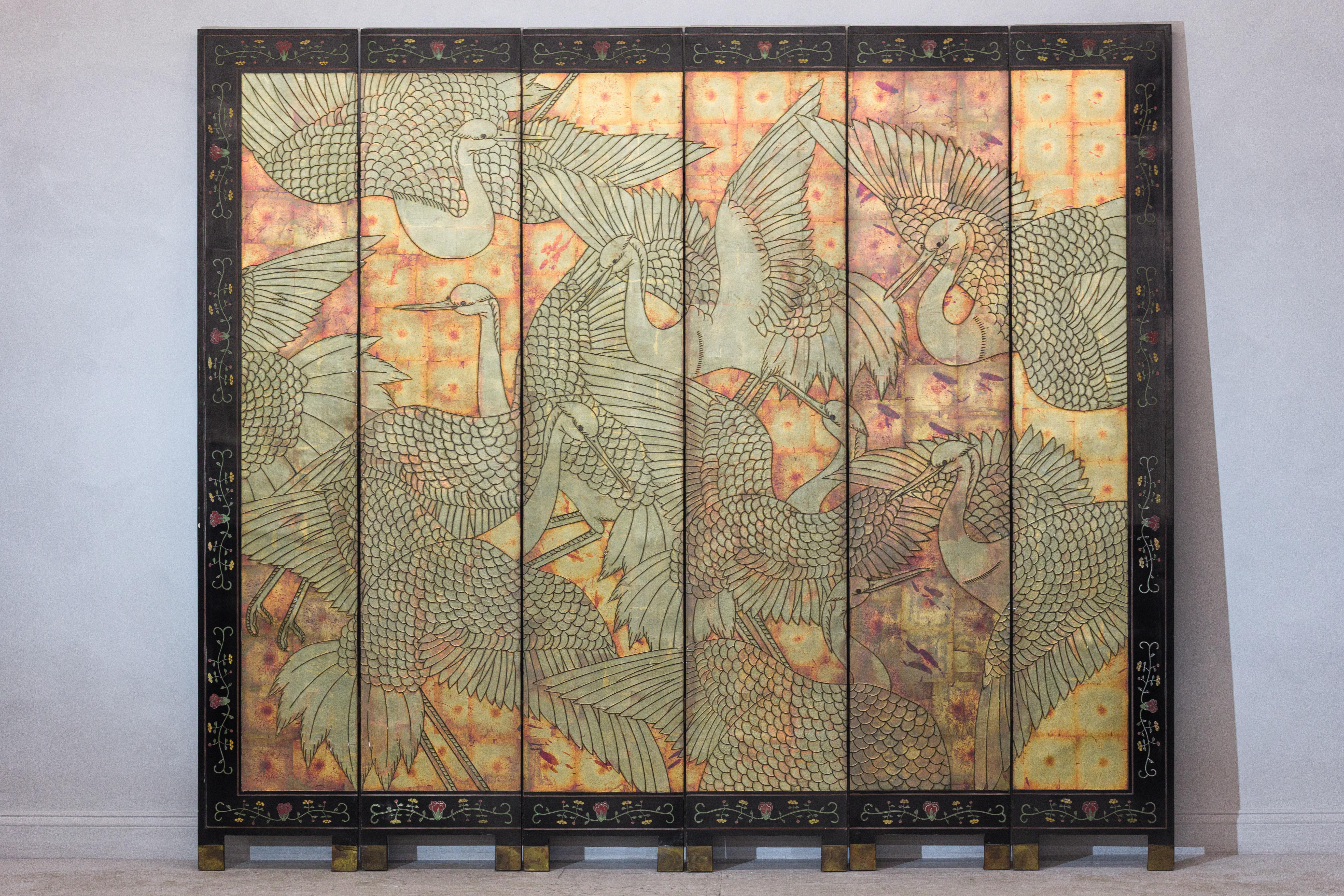 A striking six-panel Chinese Coromandel screen from the mid-20th century, adorned with intricately incised and gilded cranes in flight. Set against a warm, golden background with a timeworn patina, the composition captures the dynamic movement of