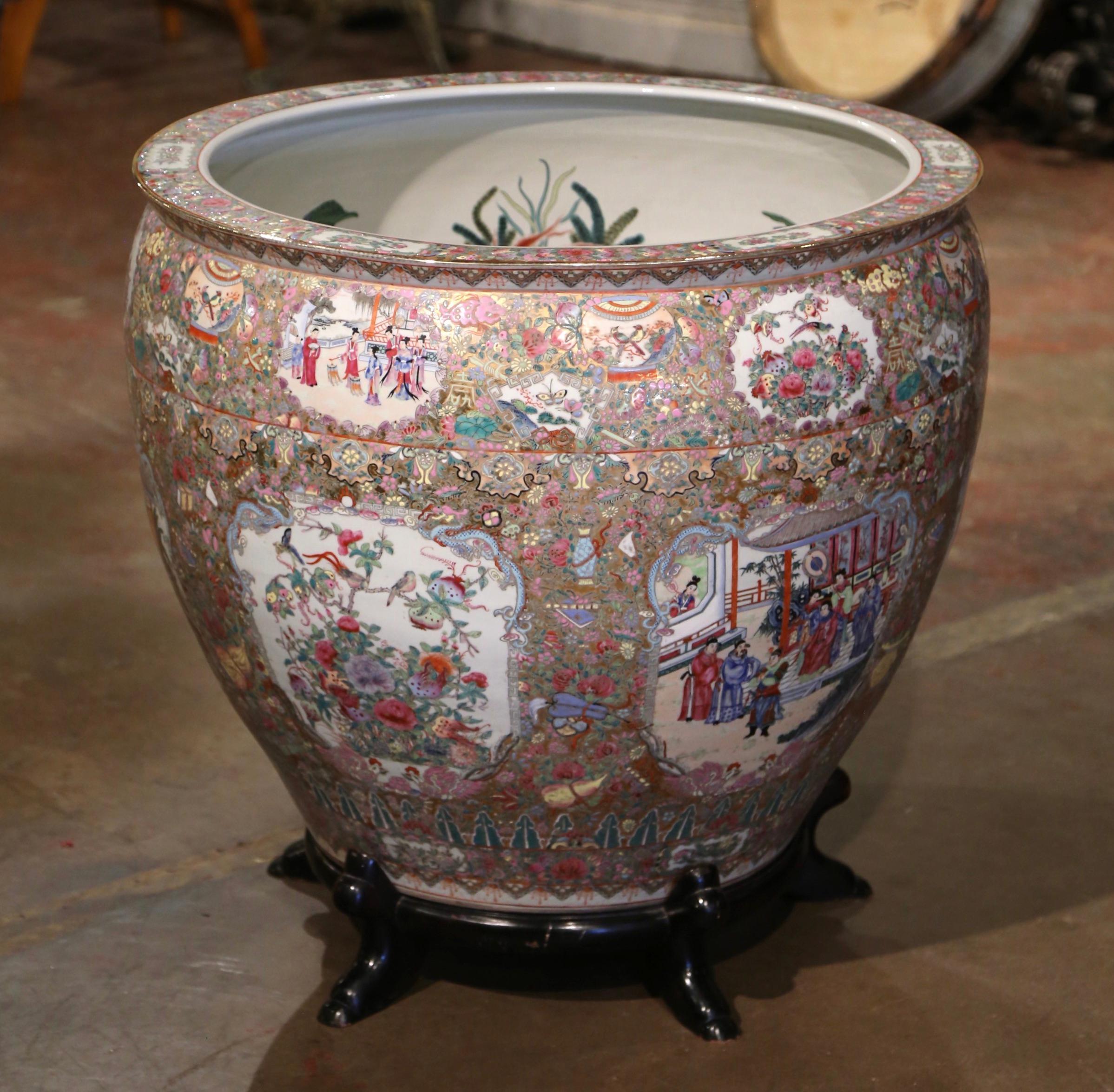 This massive and colorful vintage fishbowl was created in China, circa 1970. Round in shape and standing on a carved fruitwood base, this elegant exotic porcelain planter features hand painted medallions with Chinese figural motifs and embellished
