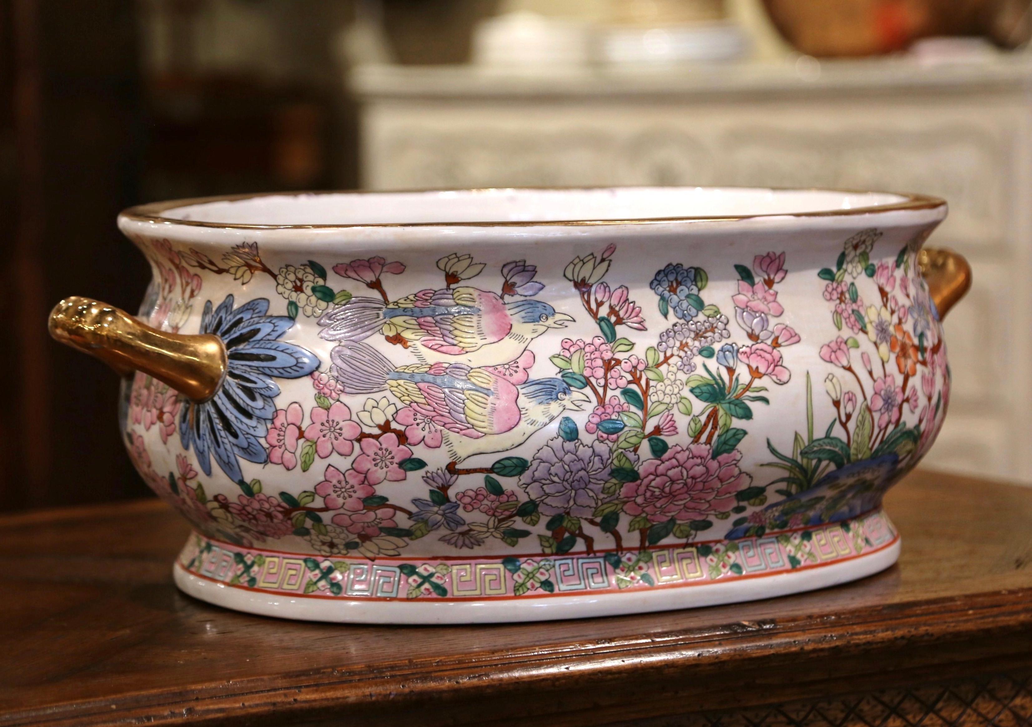 Crafted in China circa 1960 and oval in shape, the colorful porcelain basin with two side gilt handles bowl is hand painted with floral, foliage and bird motifs and embellished with a gold leaf trim around the rim. Marking on the bottom. This