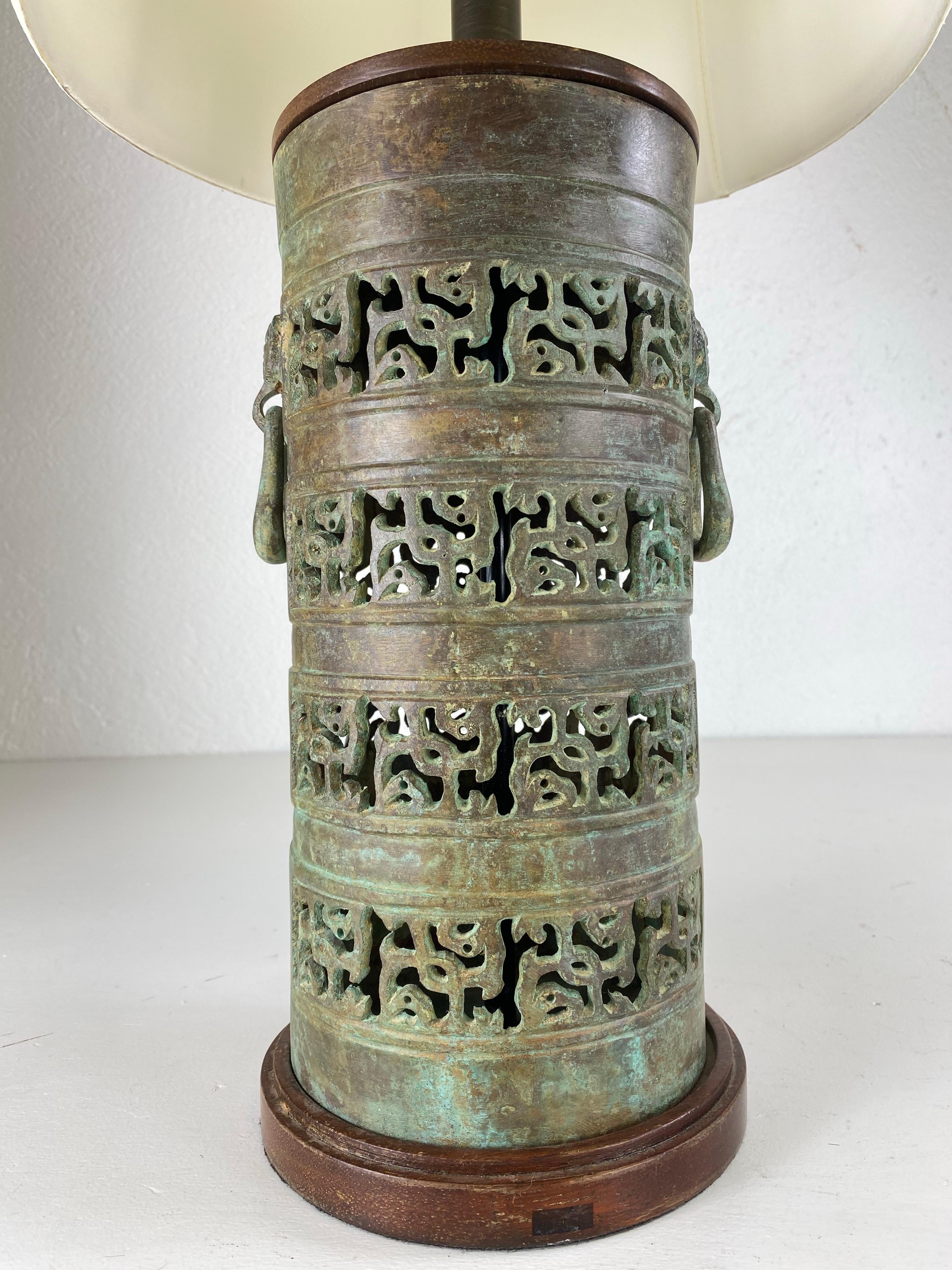 This is a mid-century vintage Mongolian inspired bronze table lamp. this lamp has a canister shape with a pierced design. The lamp has an oxidized olive green finish to the surface of the bronze. The lamp features decorative handles on either side