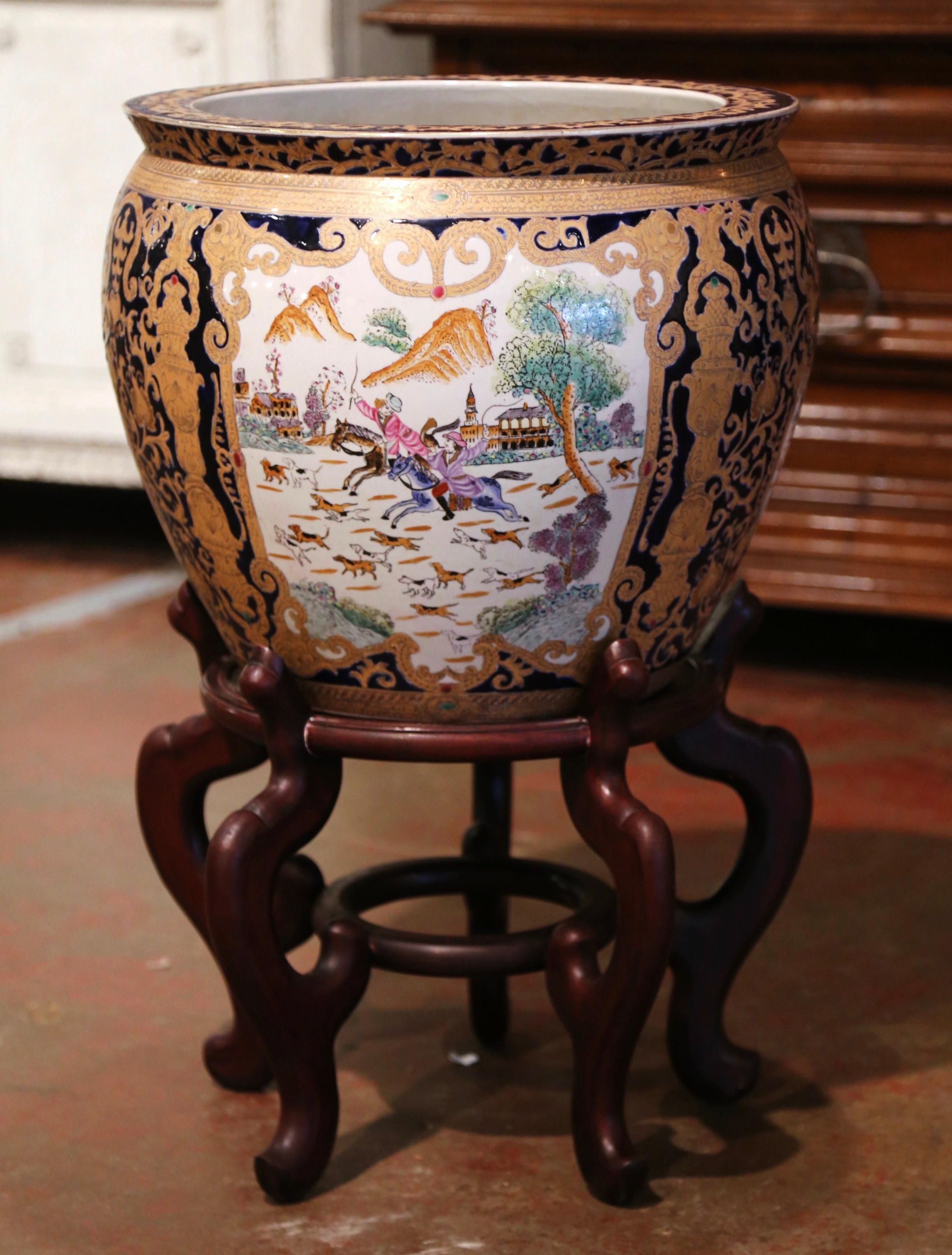 This elegant and colorful vintage fishbowl standing on a carved fruitwood base was created in China, circa 1960. Round in shape, the large, exotic porcelain planter features Classic oriental scenes with Chinese people throughout, and embellished
