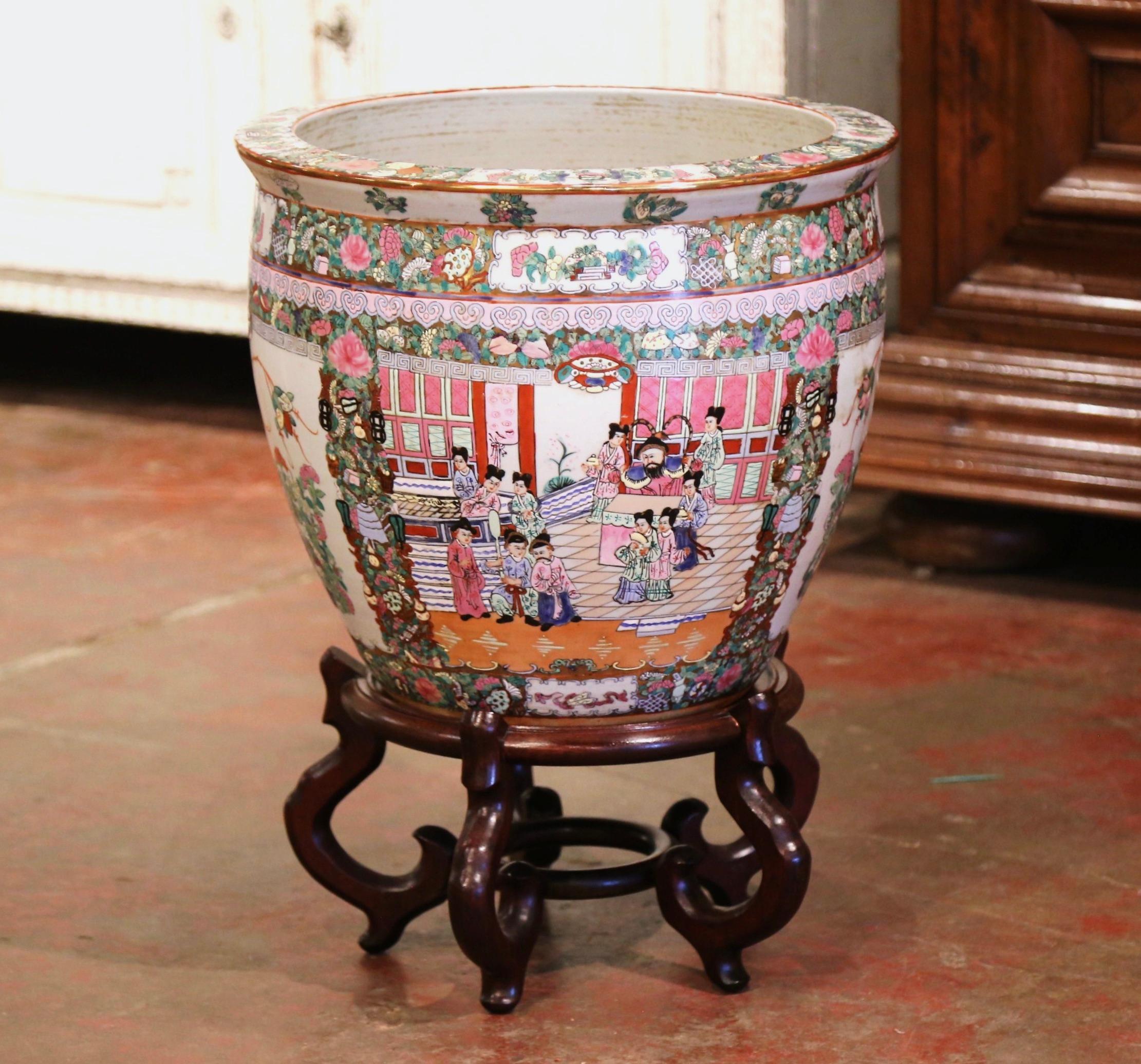 This elegant and colorful vintage fishbowl standing on a carved fruitwood base was created in China, circa 1960. Round in shape, the large, exotic porcelain planter features Classic oriental scenes with Chinese people throughout, and embellished