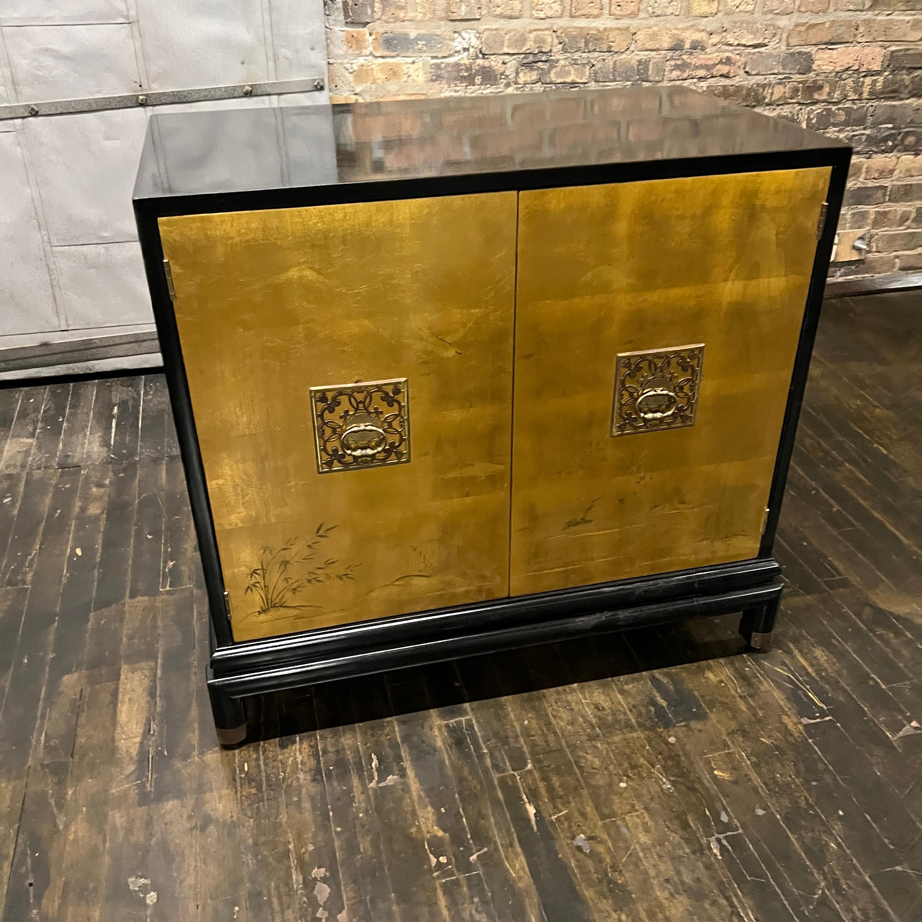 This is a stunning Chinoiserie style black lacquer cabinet with gilded (golden) doors that have images of birds, mountains and grasses painted in the gilding. There are two decorative brass door pulls. Cabinet opens to reveal one adjustable shelf.